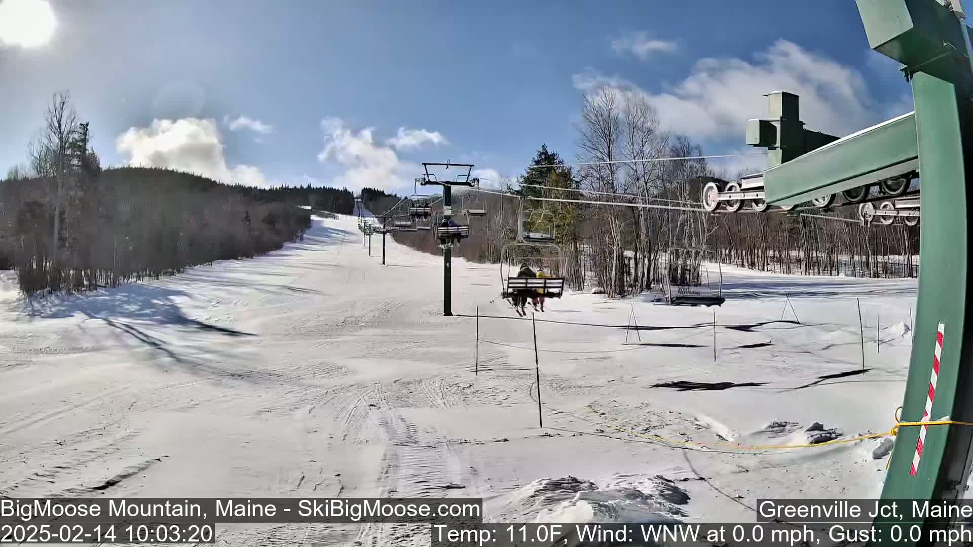 A sunny day at a ski resort shows a chairlift carrying skiers down a snow-covered slope, with trees lining the background.