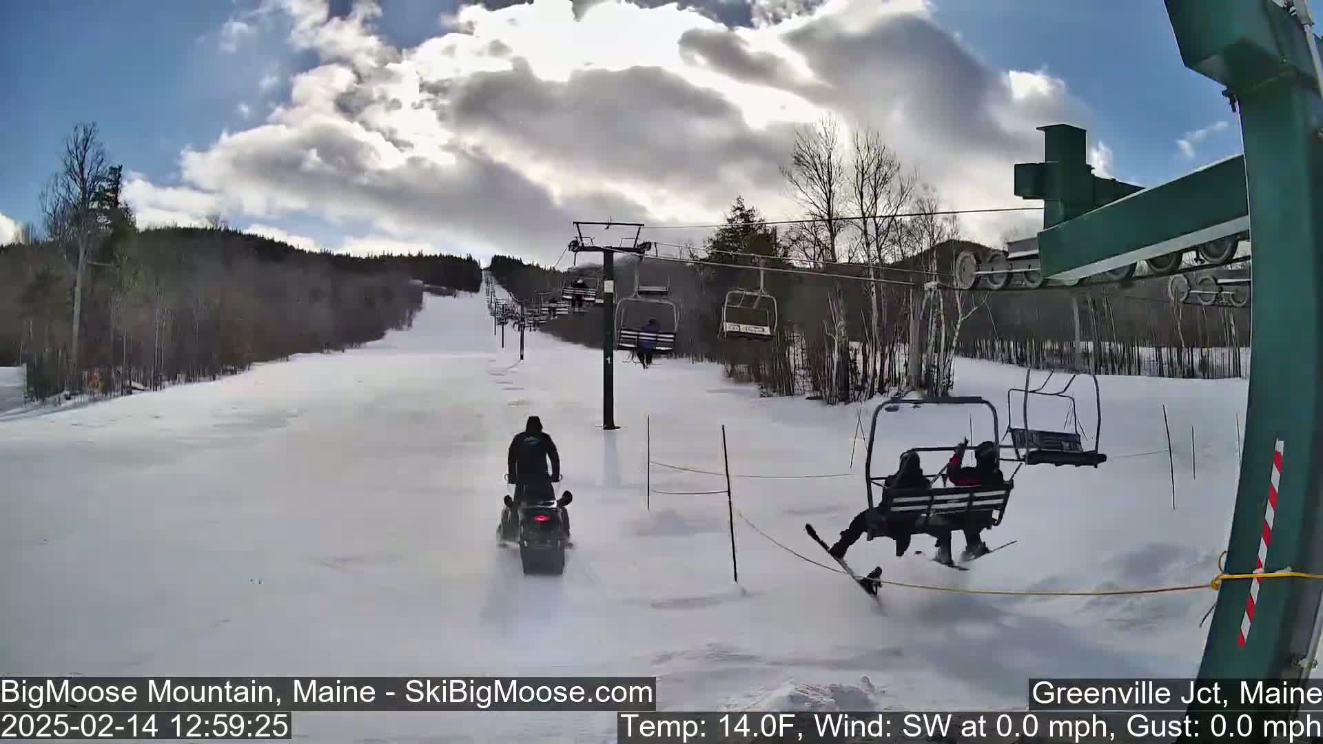 A snow-covered ski slope with a snowmobile and a chairlift, under a partly cloudy sky.