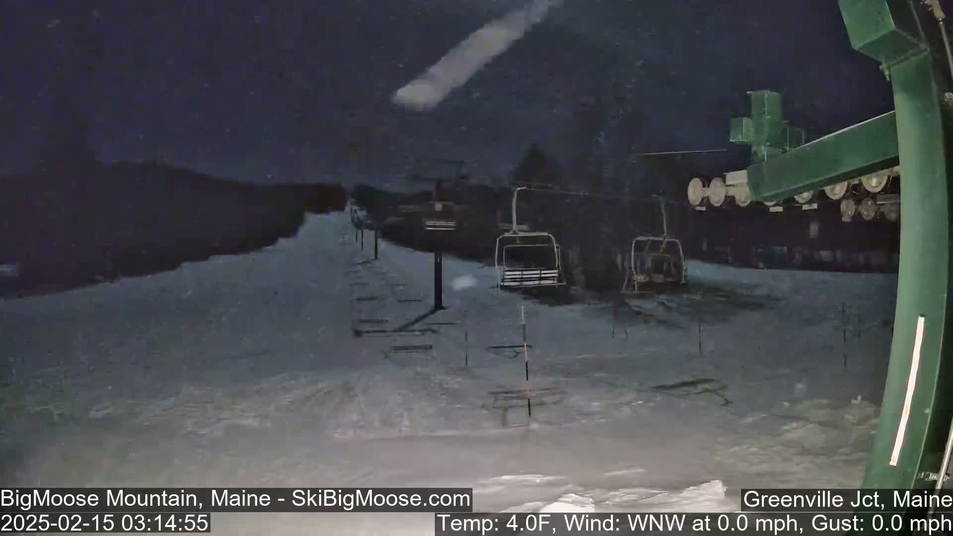 A nighttime snow scene at a ski resort shows empty chairlifts and a snow-covered slope under a dark sky with light snowfall.