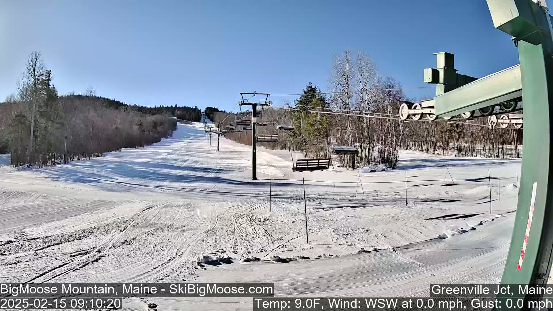 A snow-covered ski slope with a chairlift and surrounding trees under a clear, sunny sky.