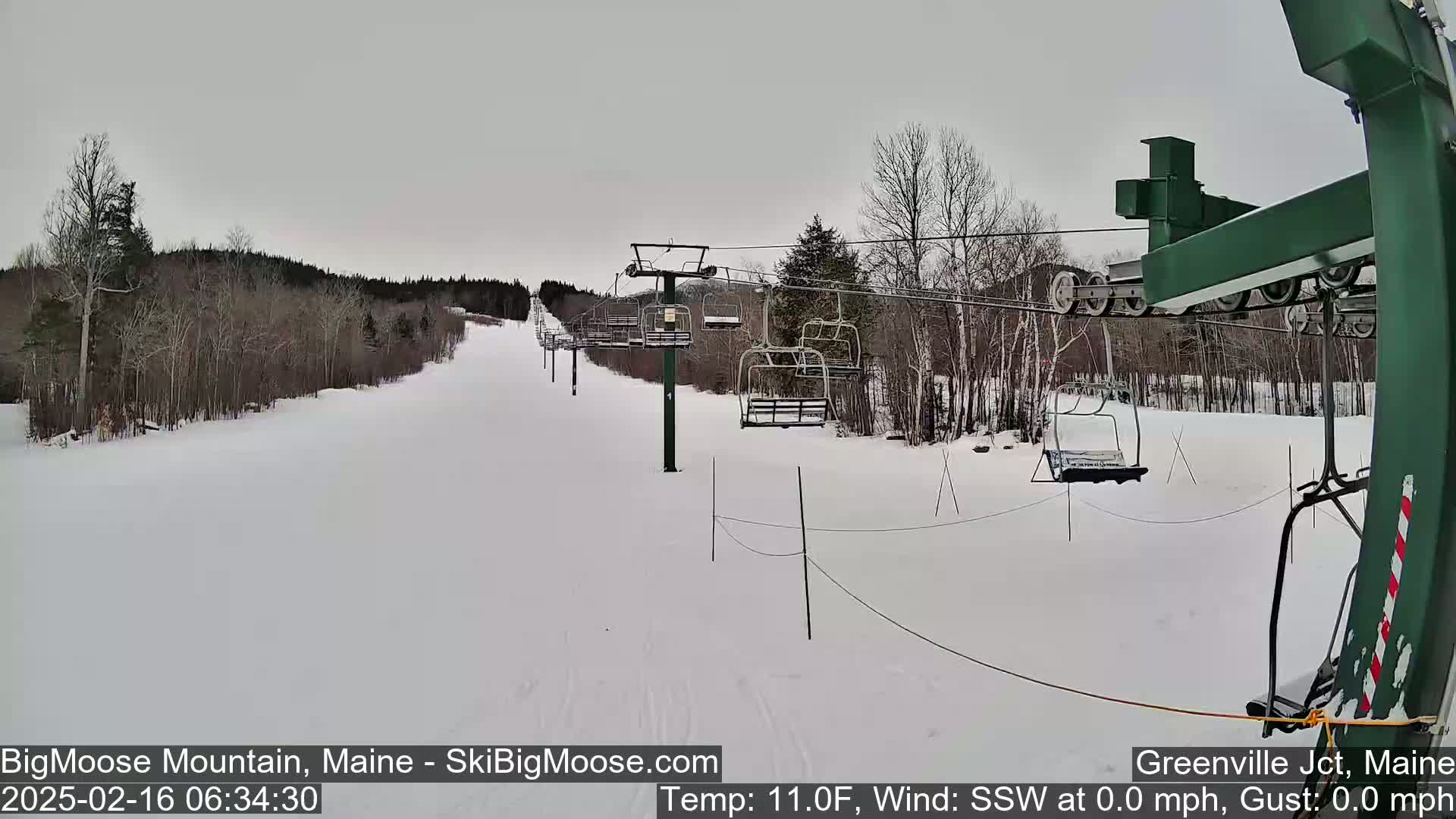 A snowy ski slope with a chairlift and sparsely wooded hills under an overcast sky.