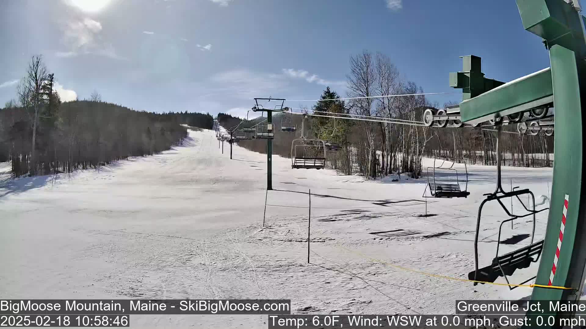 A sunny day at a snow-covered ski resort shows a chairlift and several empty chairs ascending a slope, with trees lining the sides.