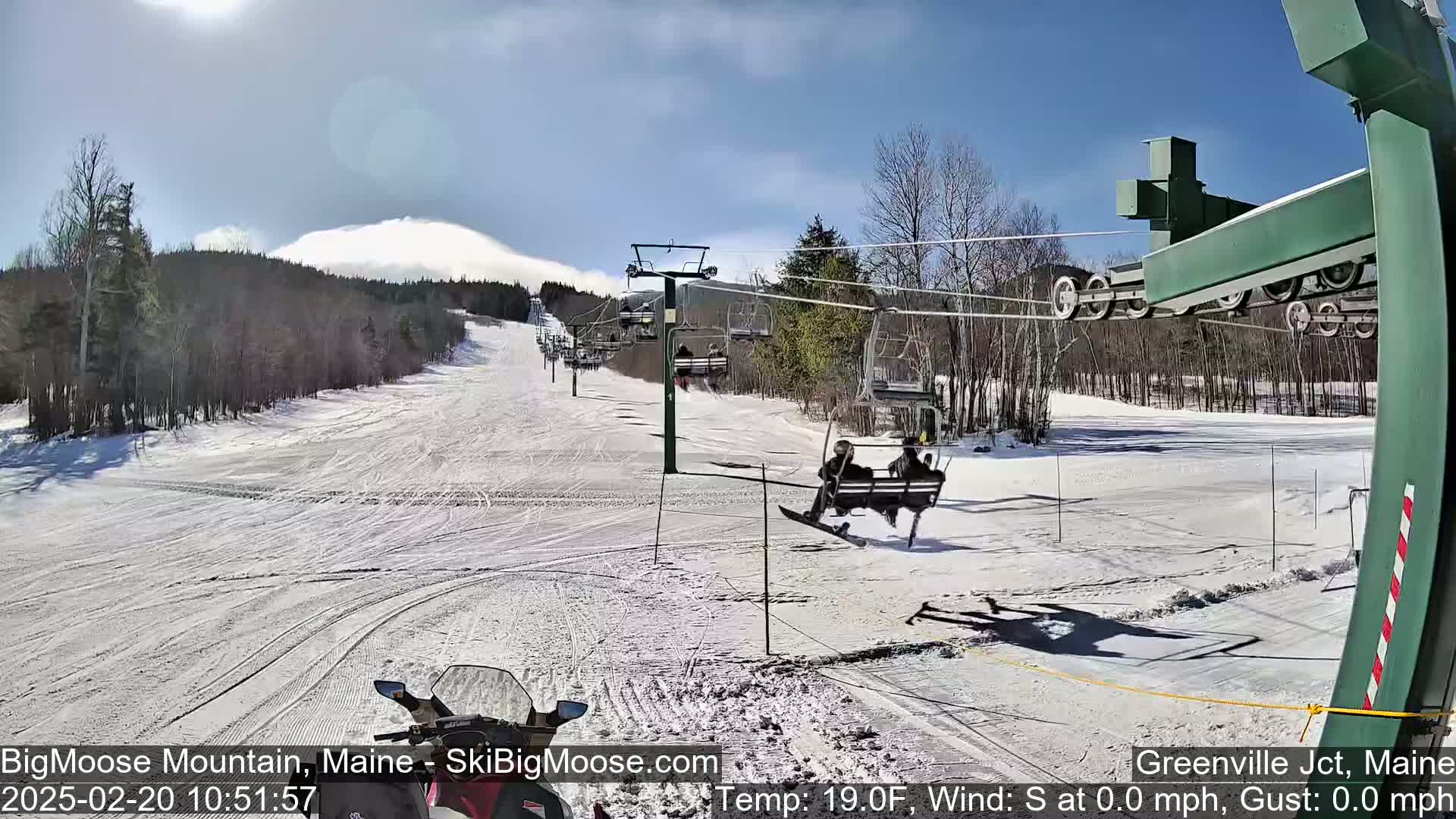 A snow-covered ski slope with a chairlift and skiers, under a mostly sunny sky.