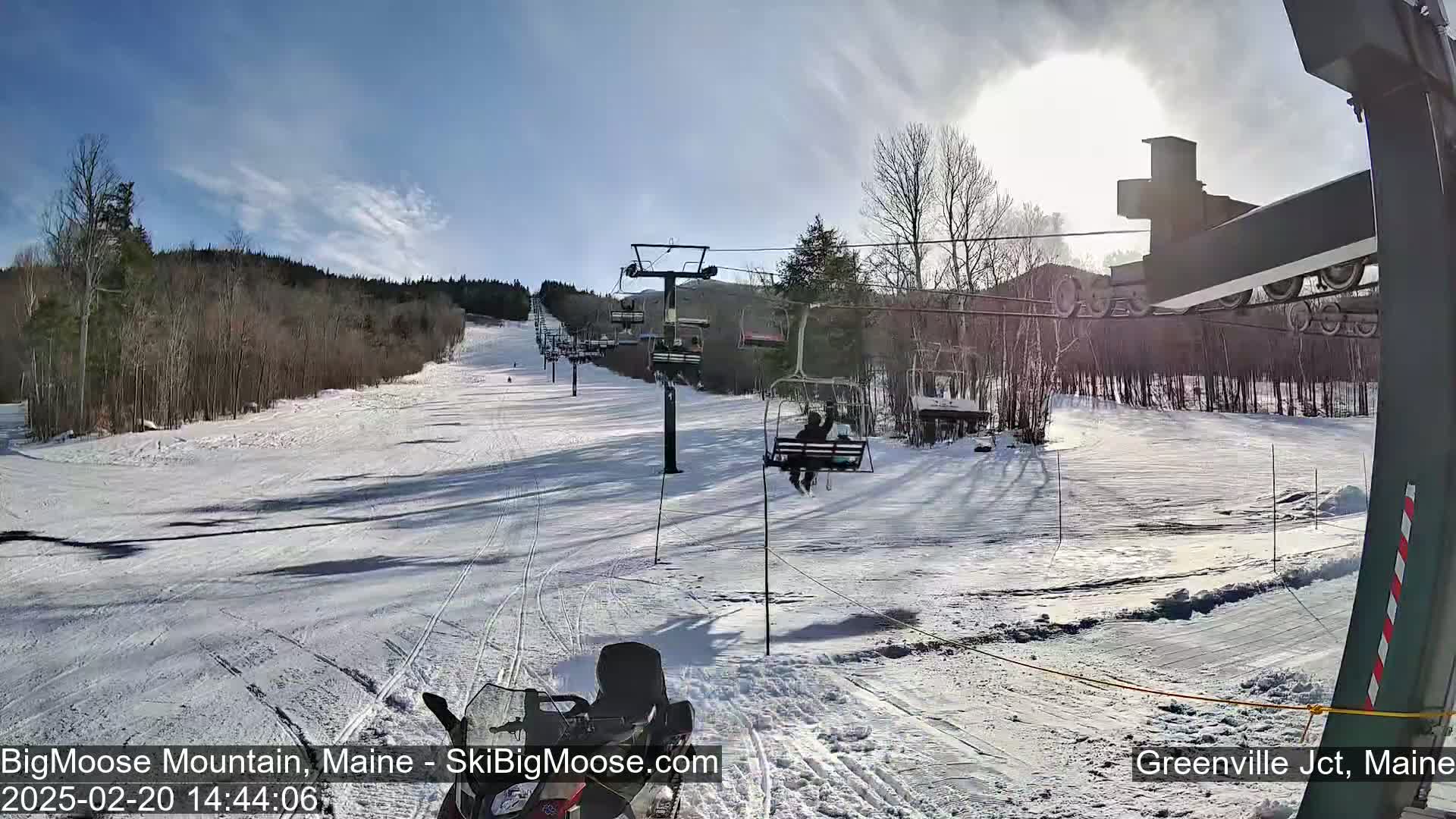 A sunny day at a snow-covered ski resort shows a chairlift carrying skiers and snowboarders up a slope, with a snowmobile visible in the foreground.