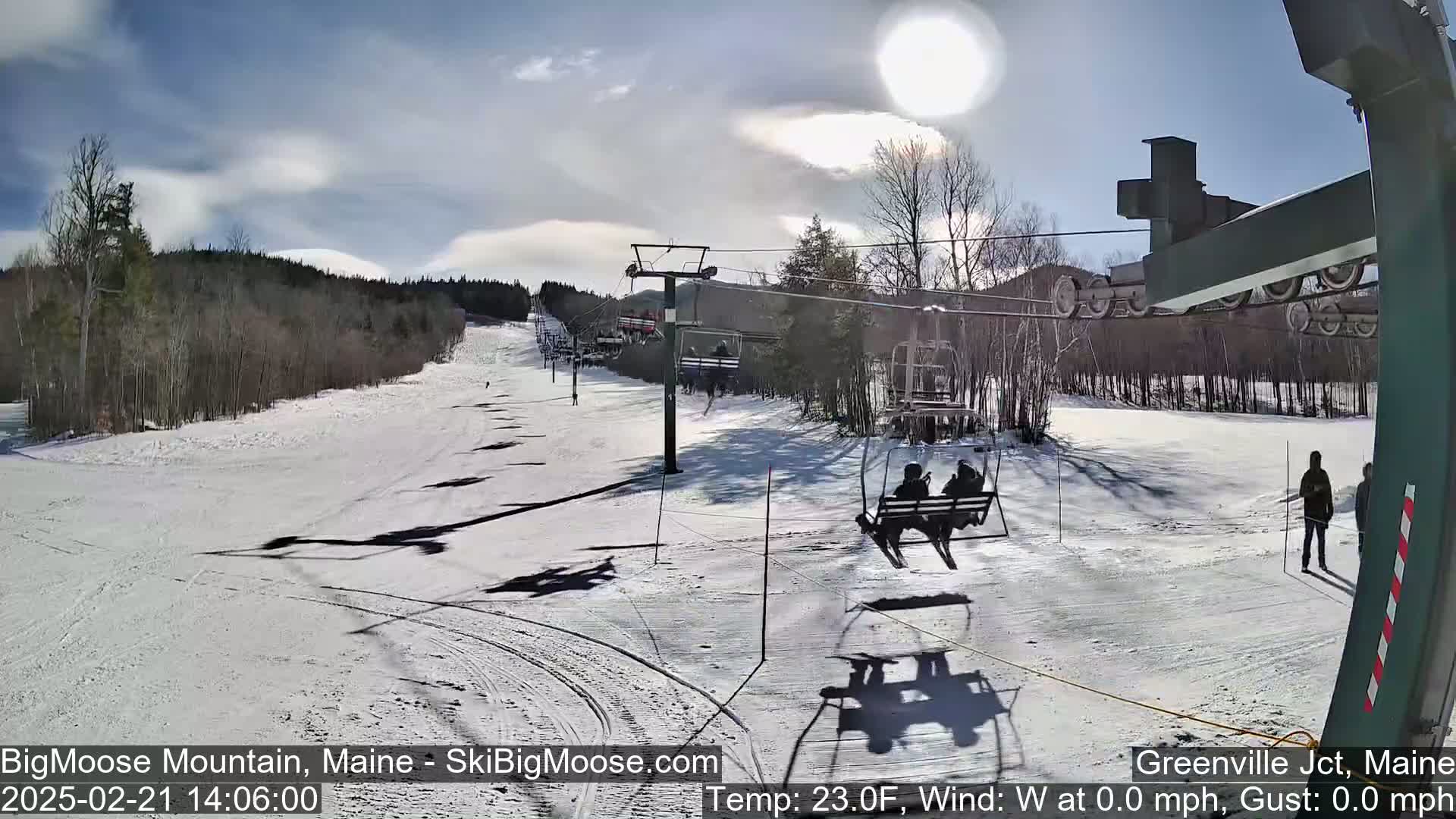 A sunny, snow-covered ski slope with a chairlift carrying skiers, and several people standing near the bottom of the lift.