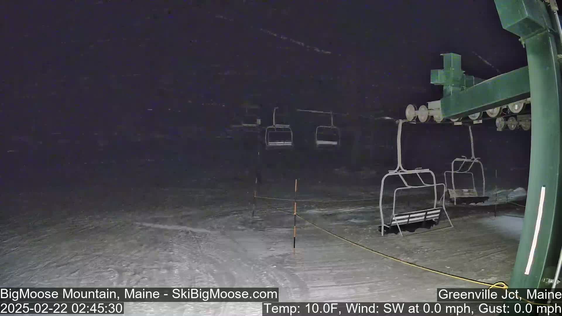 A nighttime view of a ski lift with empty chairs on a snowy slope under lightly falling snow.