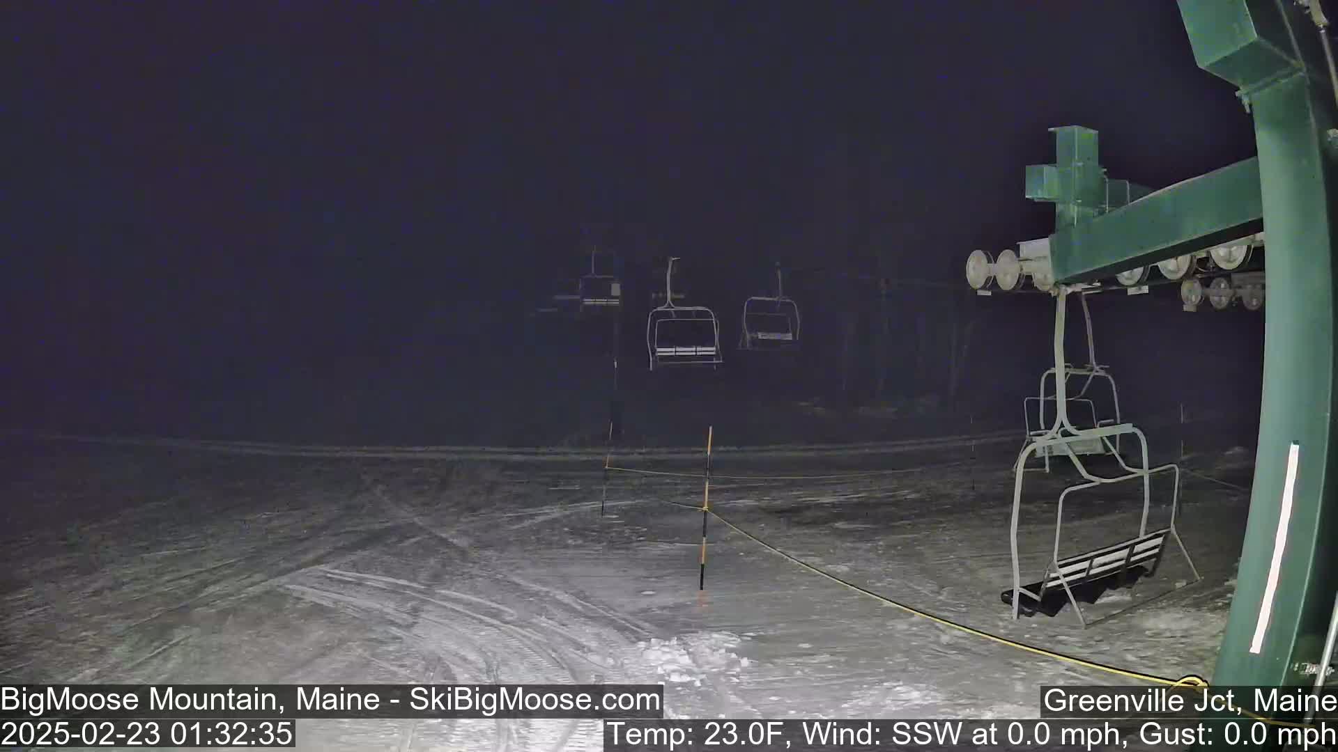 A nighttime view of a ski lift with empty chairlifts on a snow-covered surface under dark conditions.