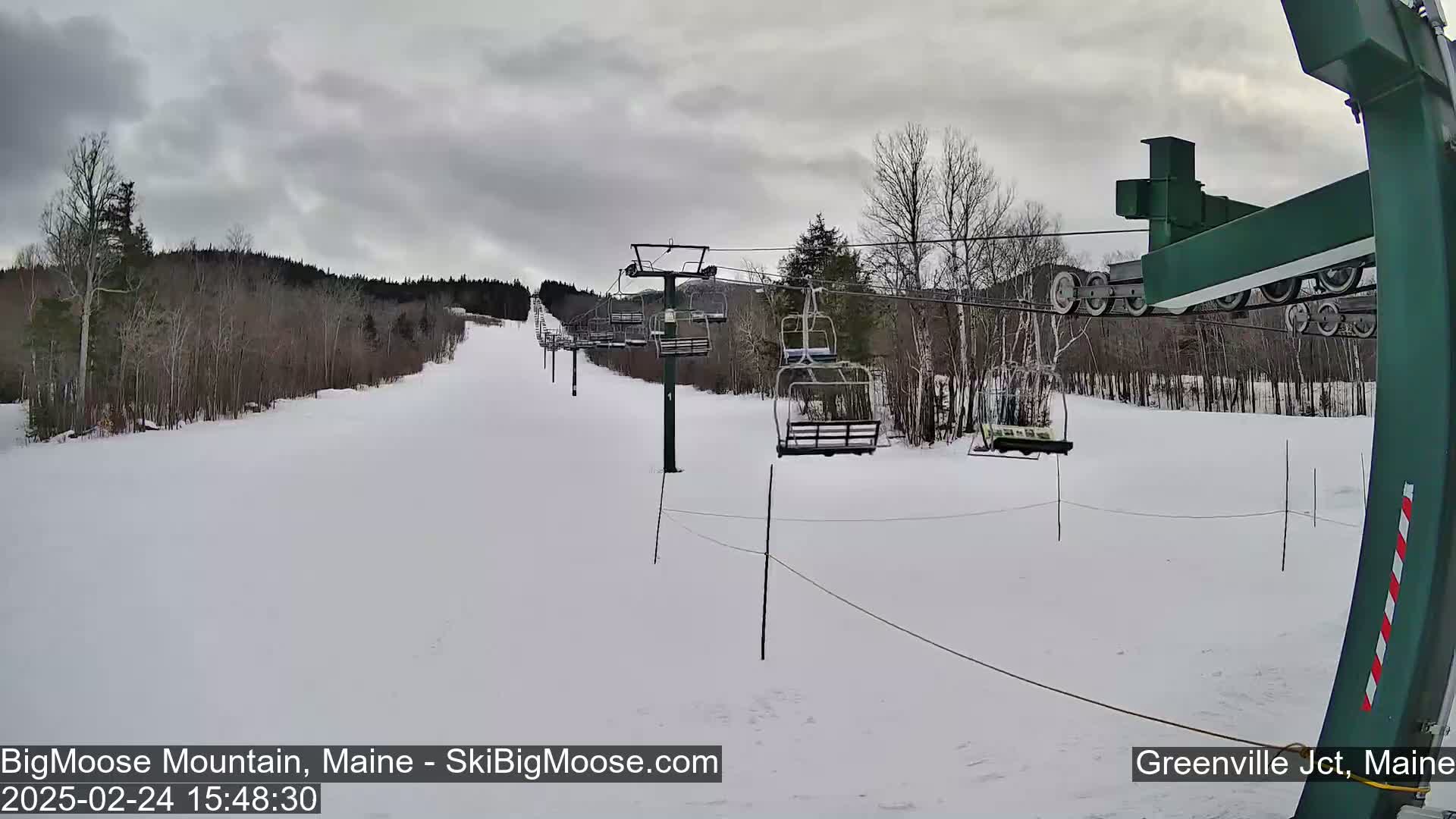 A snow-covered ski slope with a chairlift and sparsely wooded area under an overcast sky.