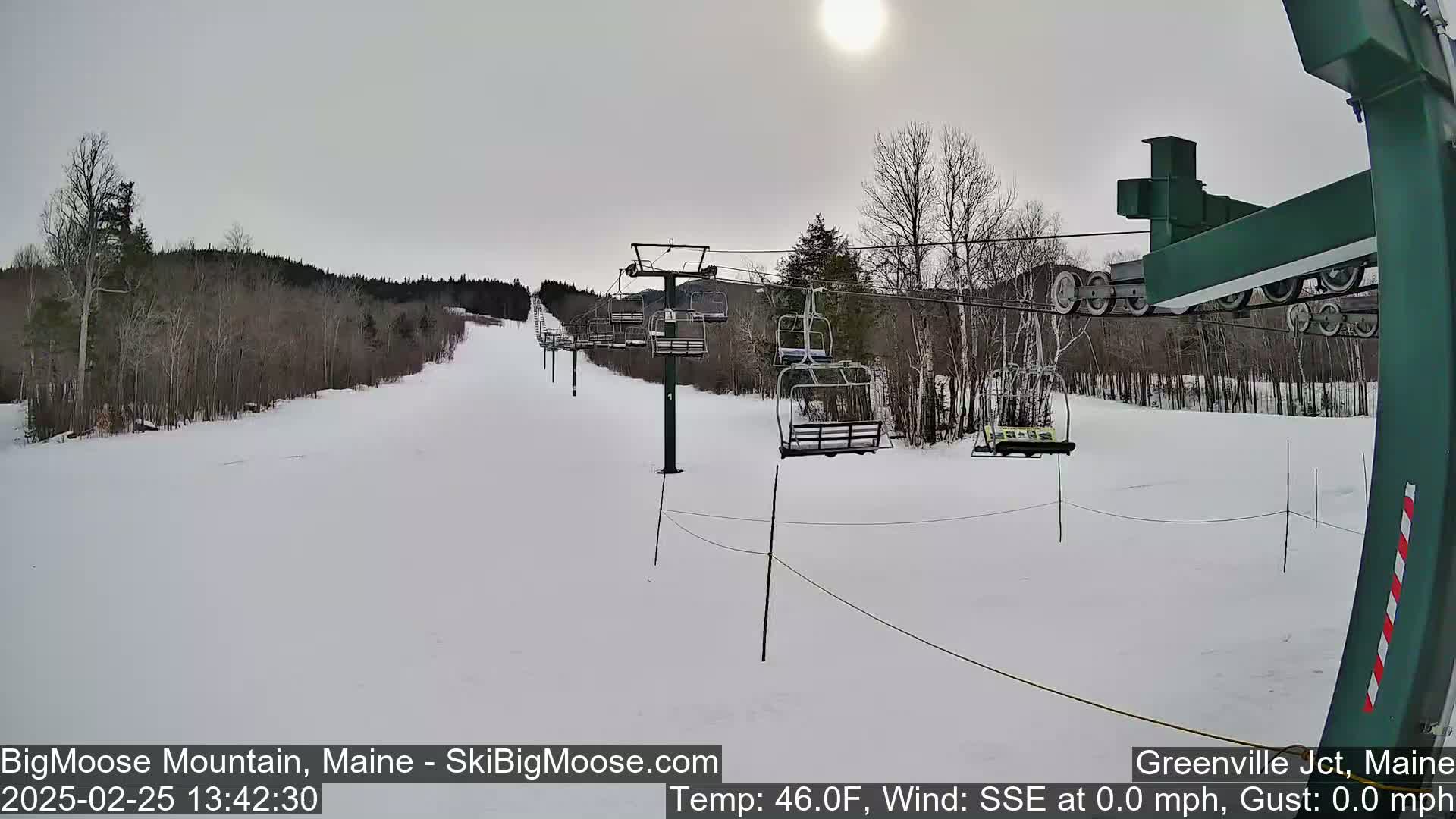 A mostly sunny, snow-covered ski slope with a chairlift and sparsely wooded hills in the background.