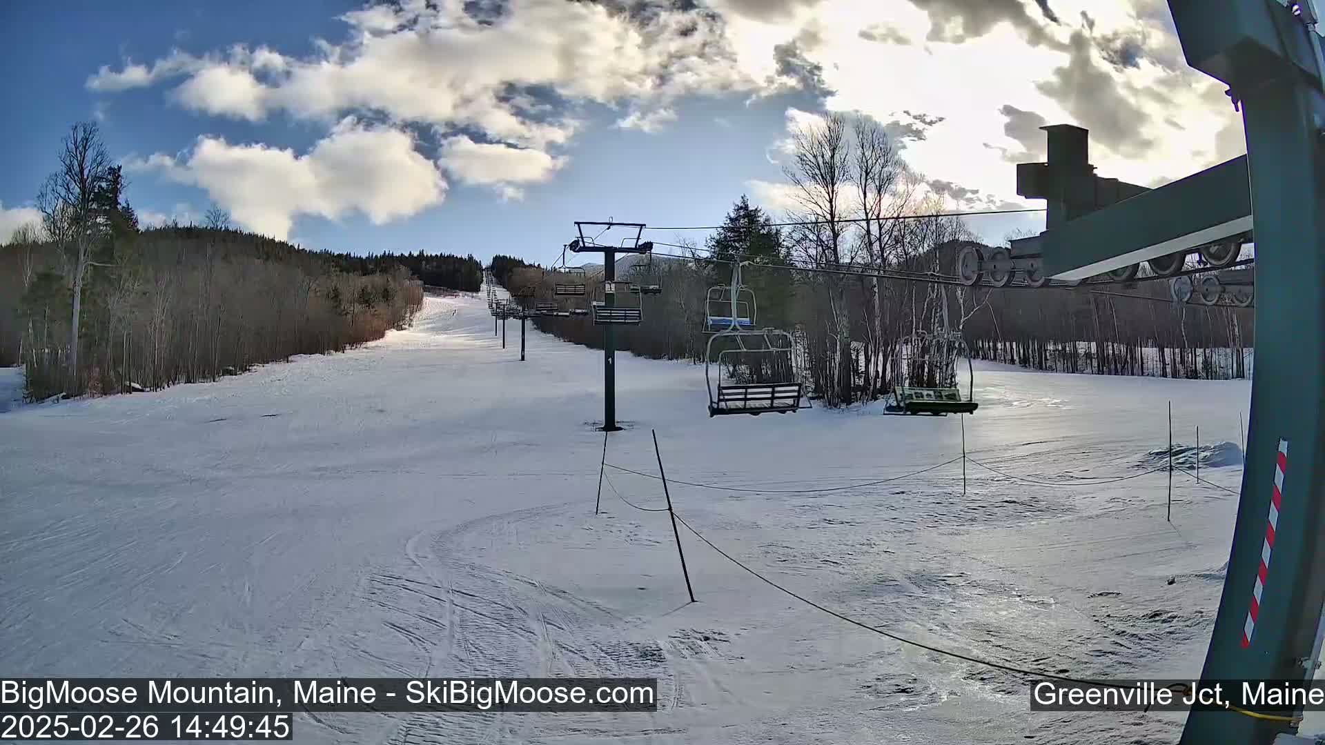 A mostly sunny day at a snow-covered ski resort shows a chairlift ascending a slope lined with bare trees.