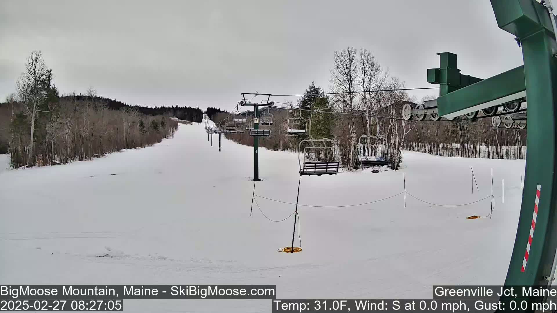 A snow-covered ski slope with a chairlift and sparsely wooded hills under a gray sky.