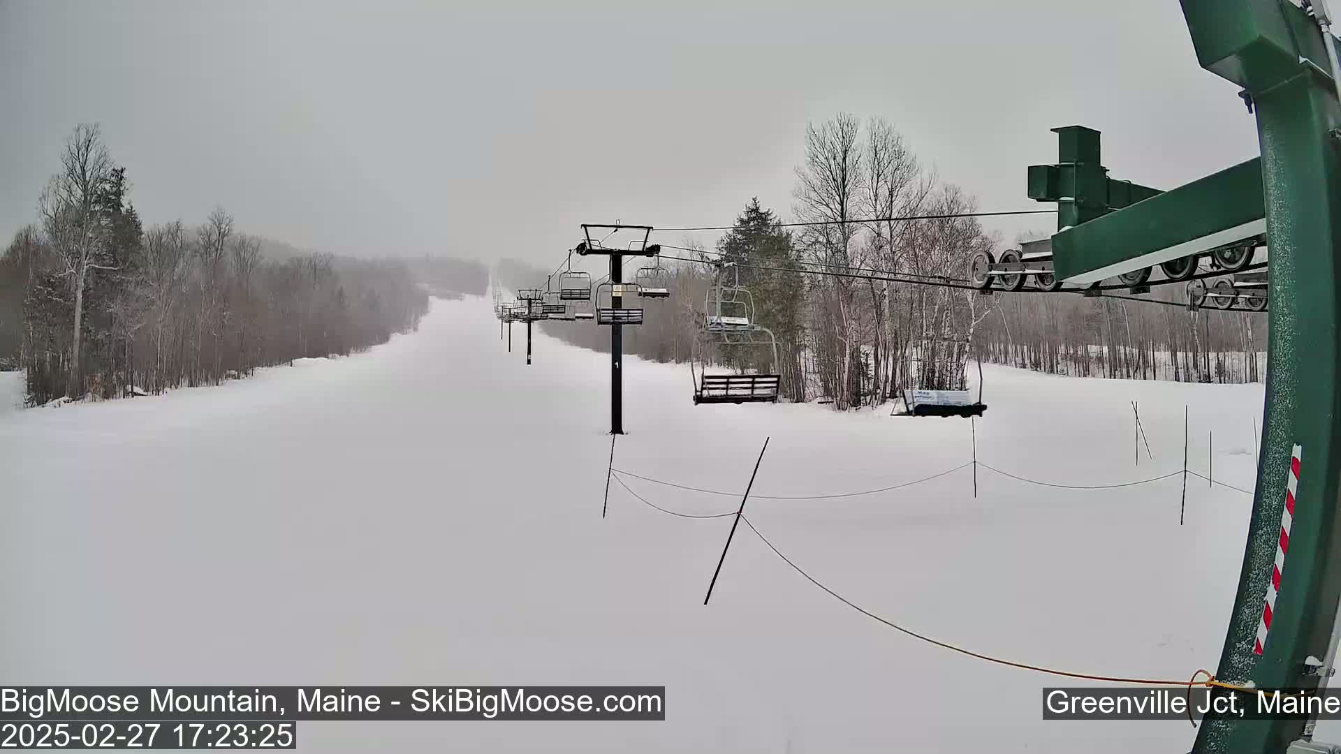 A snow-covered ski slope with a chairlift and sparsely wooded areas under an overcast sky.