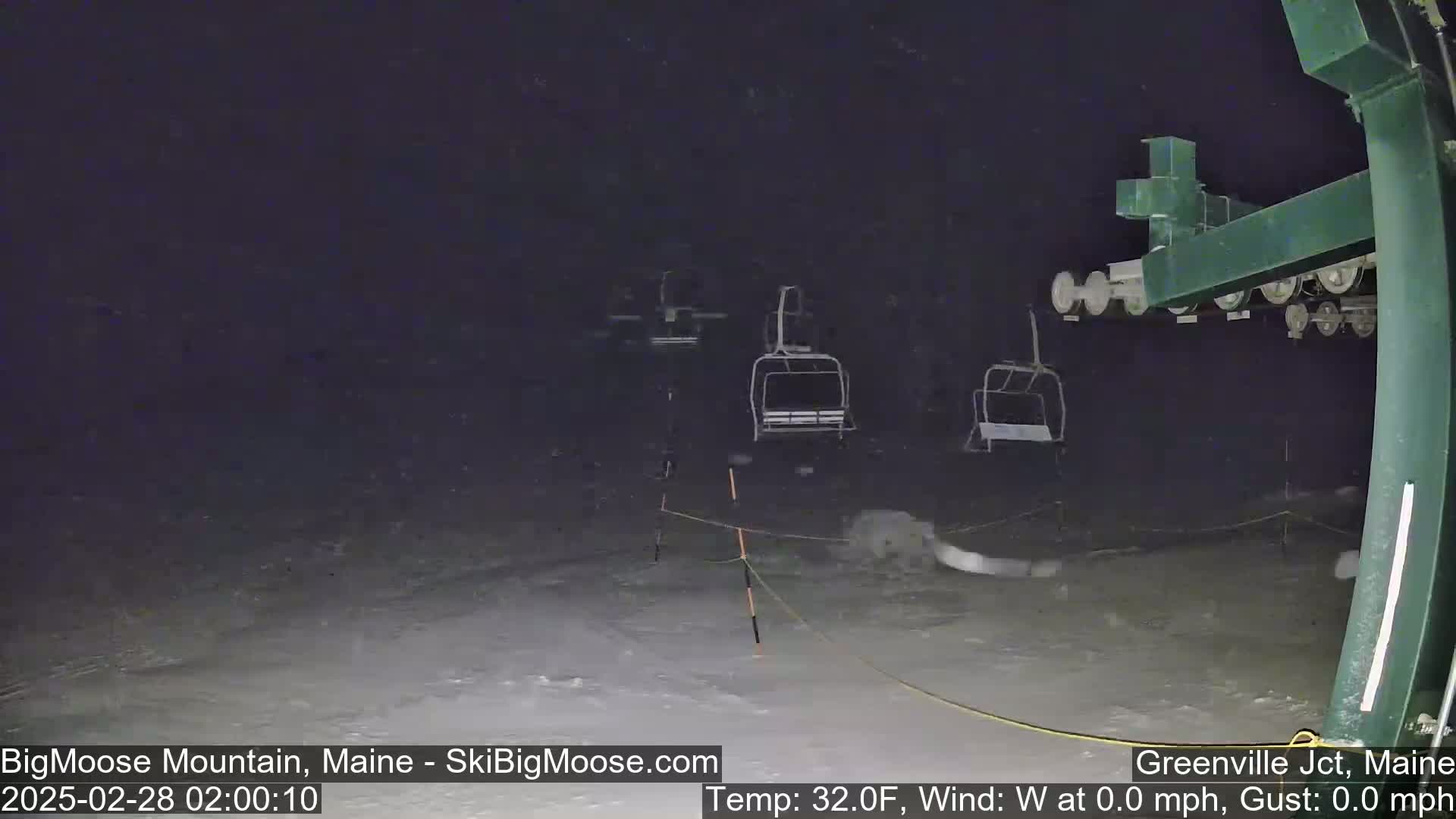 A nighttime view of a snow-covered ski lift with several empty chairs under a dark sky with light snowfall.