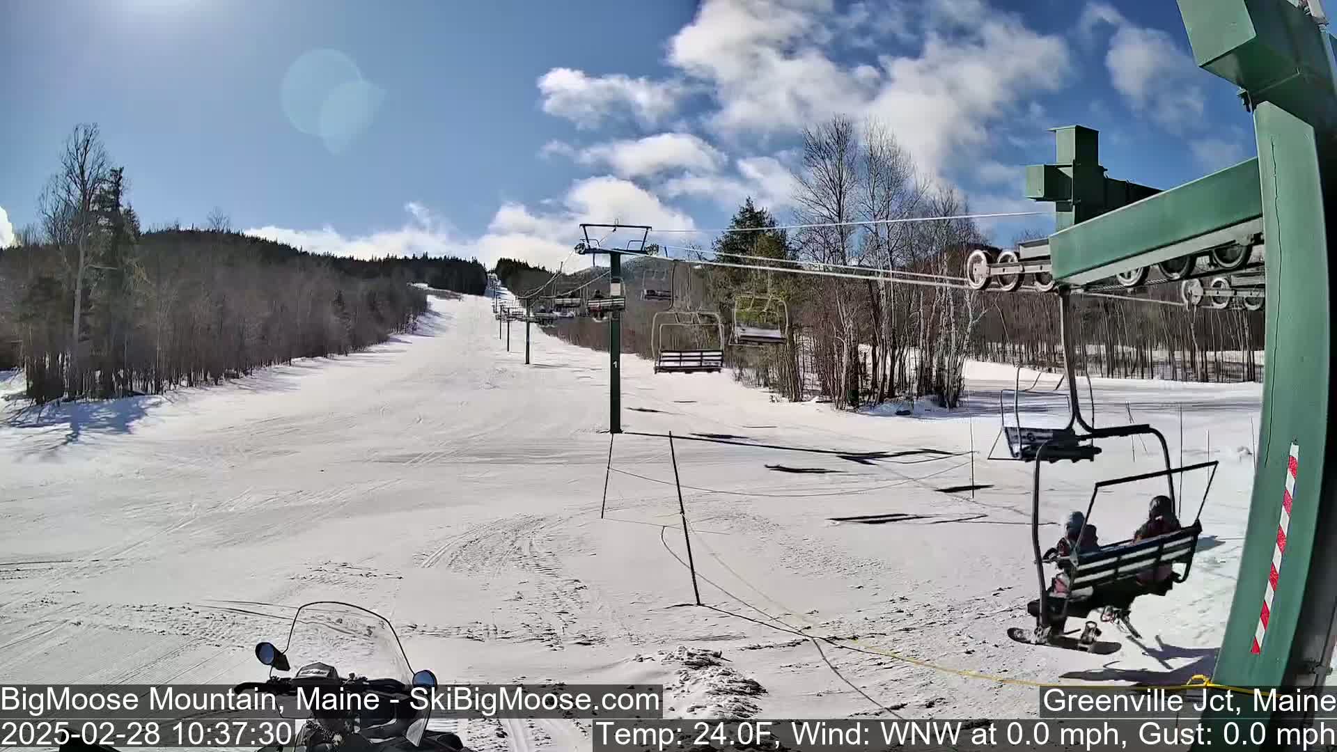 A sunny day at a snow-covered ski resort shows a chairlift carrying two people, with a snowmobile partially visible in the foreground.