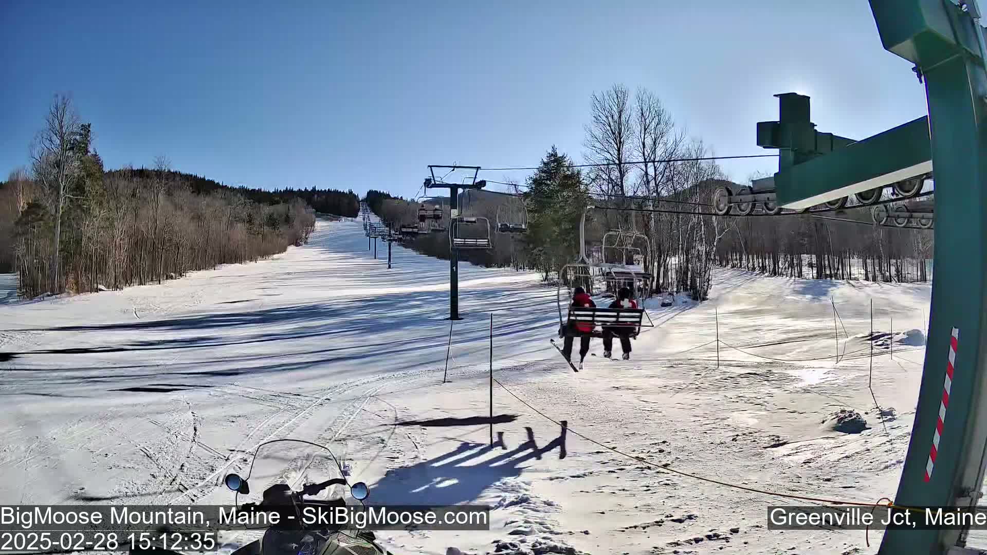 A sunny day at a snow-covered ski resort shows a chairlift carrying two skiers, with a snowmobile visible in the foreground.