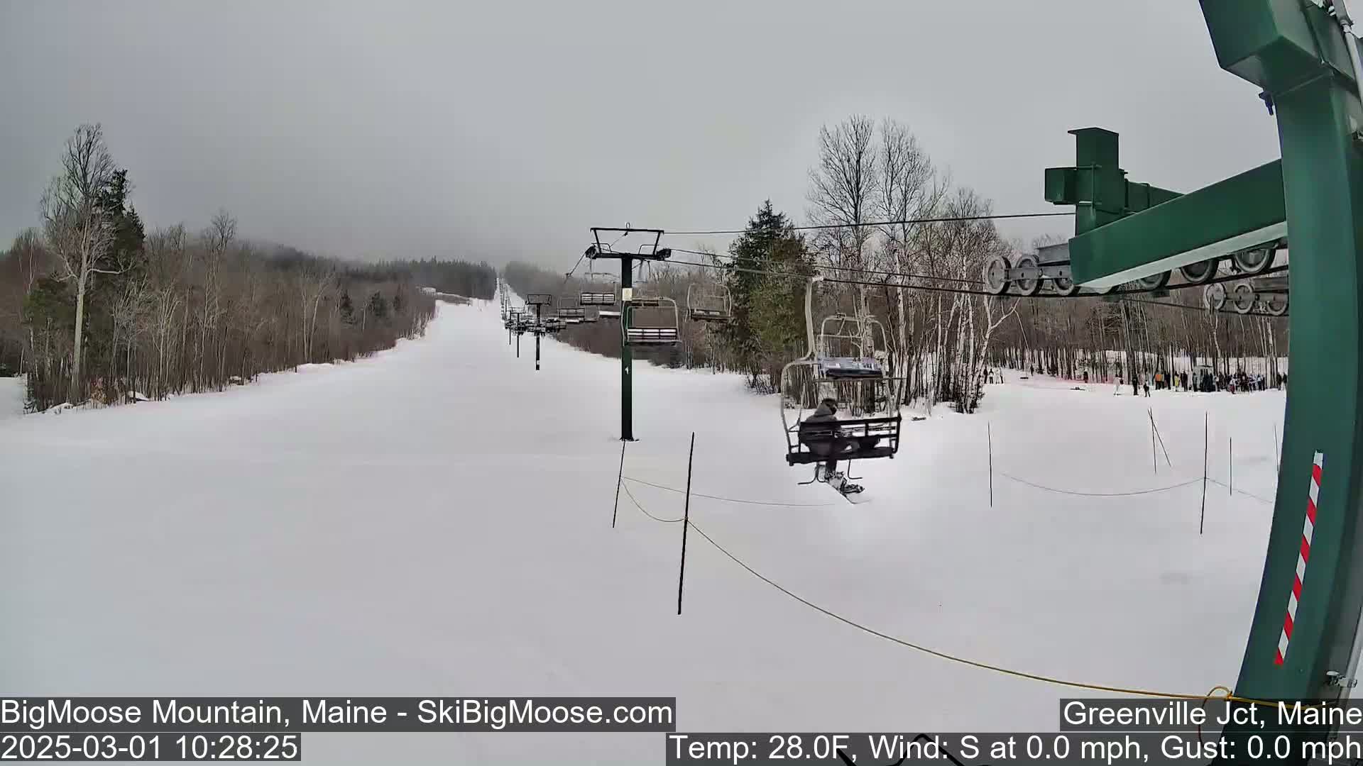 A snow-covered ski slope with a chairlift and several skiers visible on a cloudy day.