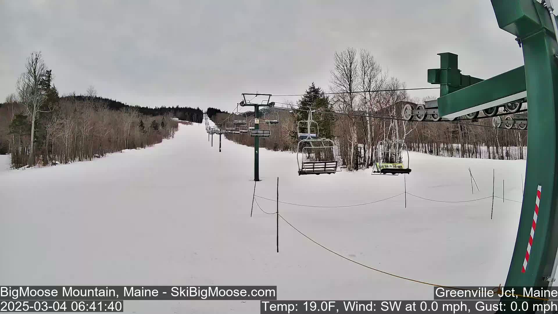 A snow-covered ski slope with a chairlift and bare trees under an overcast sky.