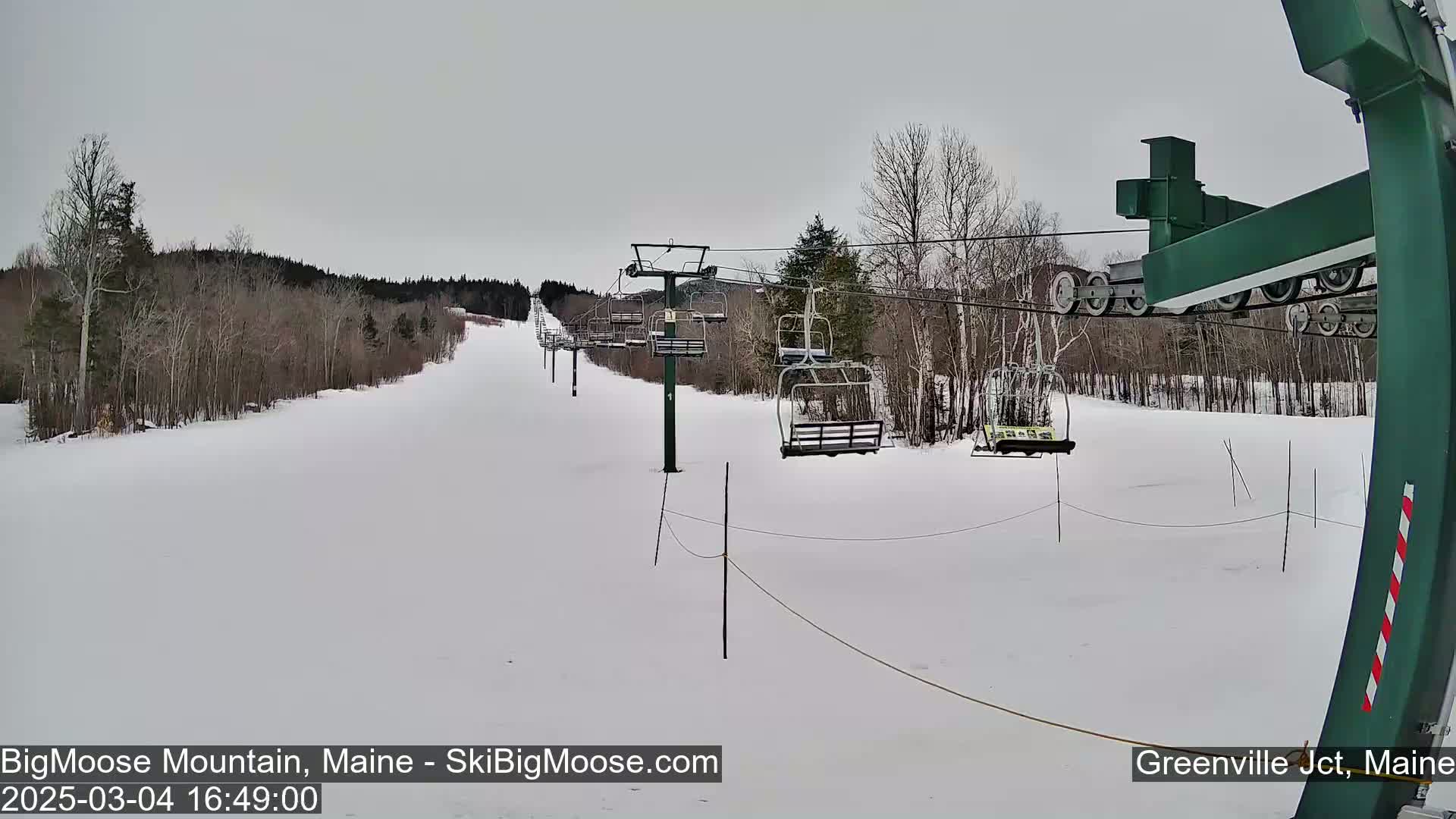 A snow-covered ski slope with a chairlift and bare trees under an overcast sky.