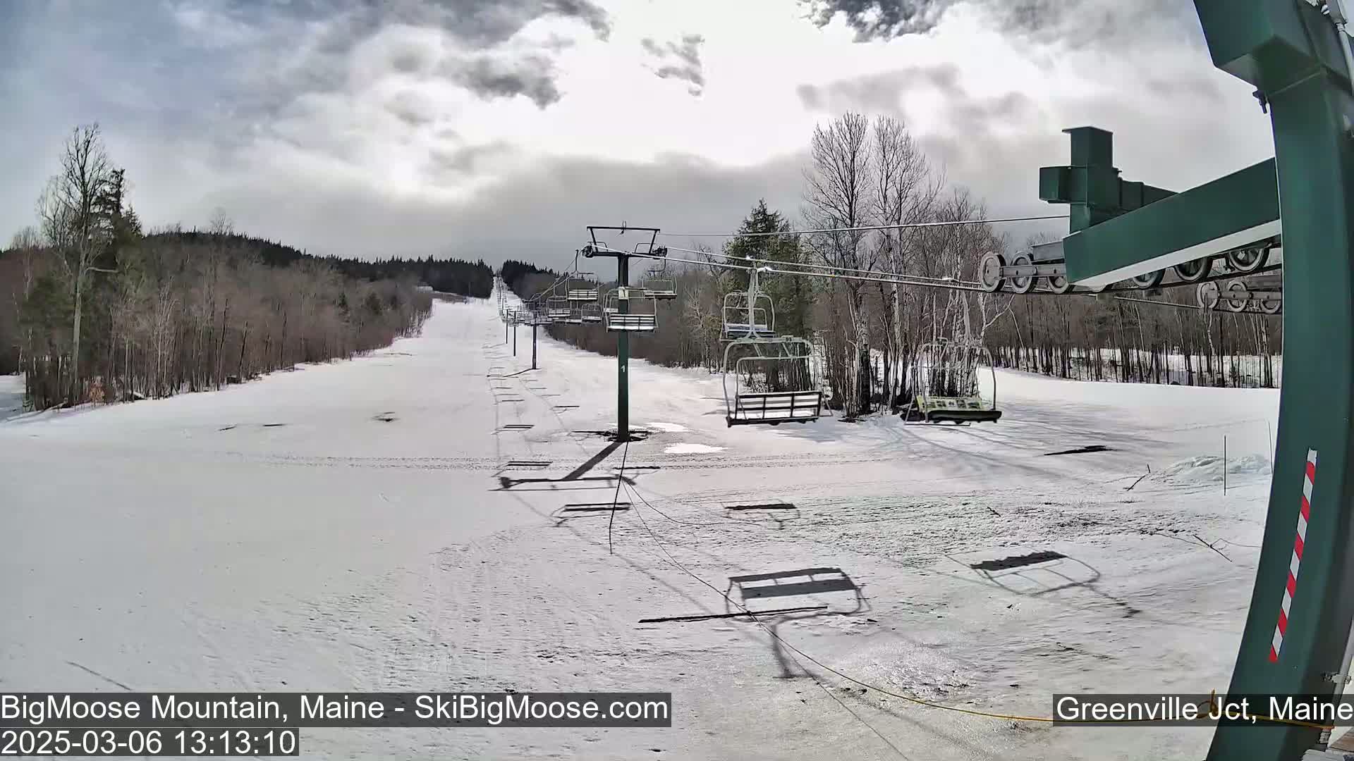 A mostly cloudy day at a ski resort shows a snow-covered ski slope with an empty chairlift and bare trees in the background.