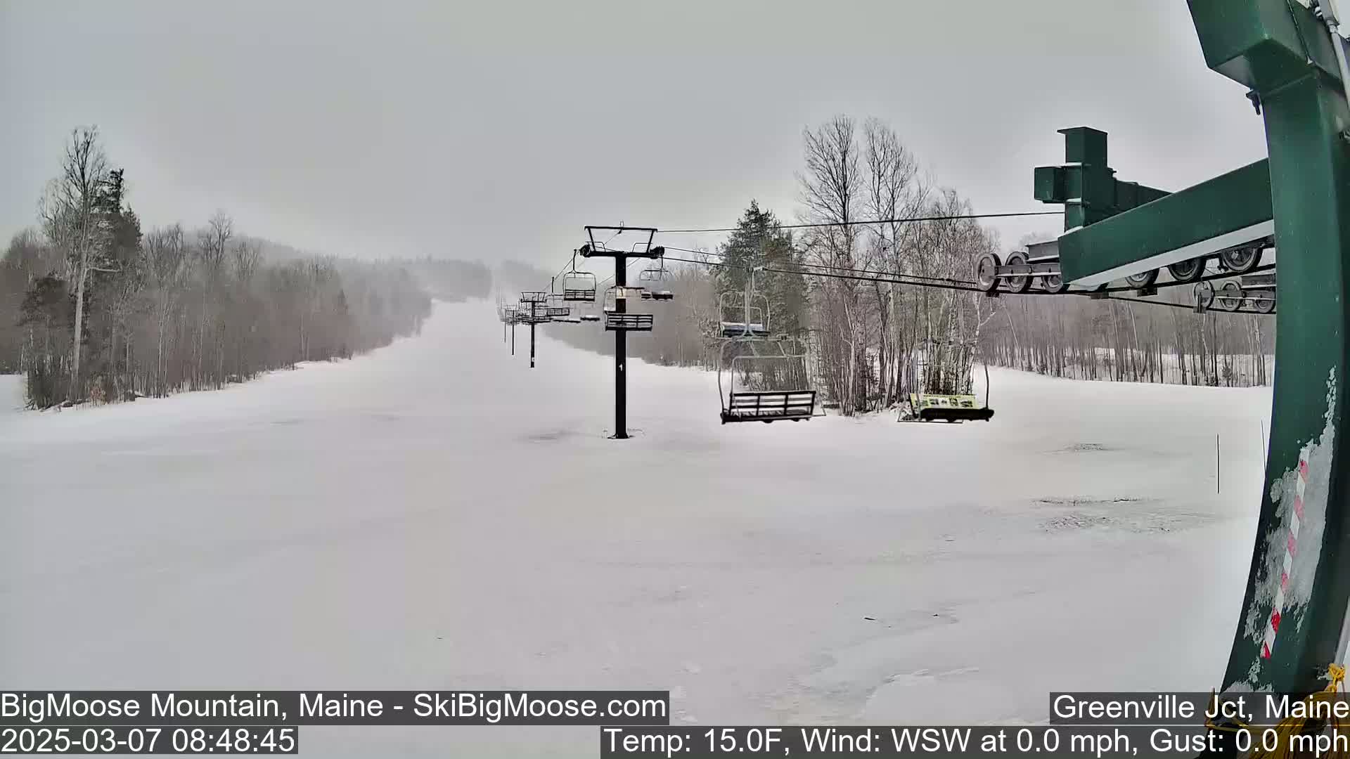 A snow-covered ski slope with a chairlift and sparsely wooded areas in overcast conditions.
