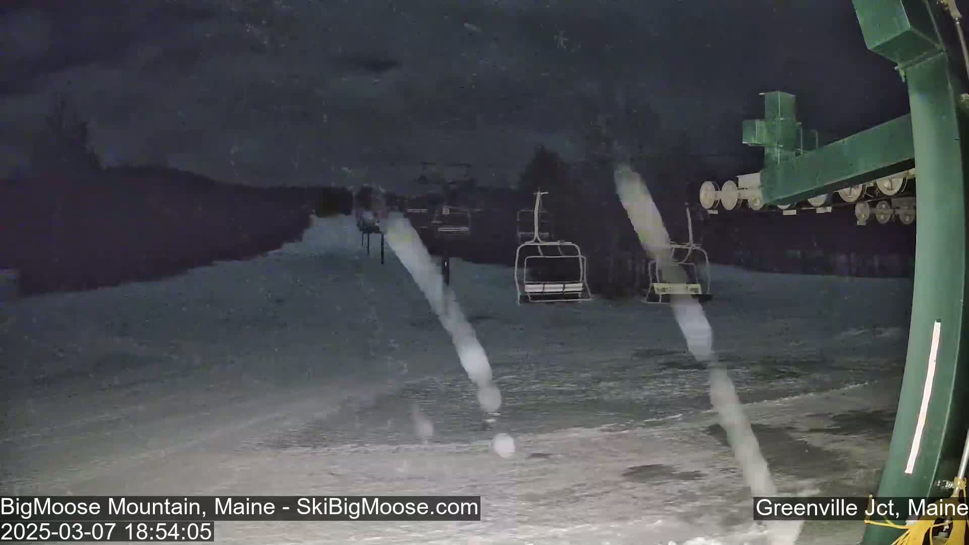A nighttime view of a snow-covered ski slope with several empty chairlifts under a dark, snowy sky.
