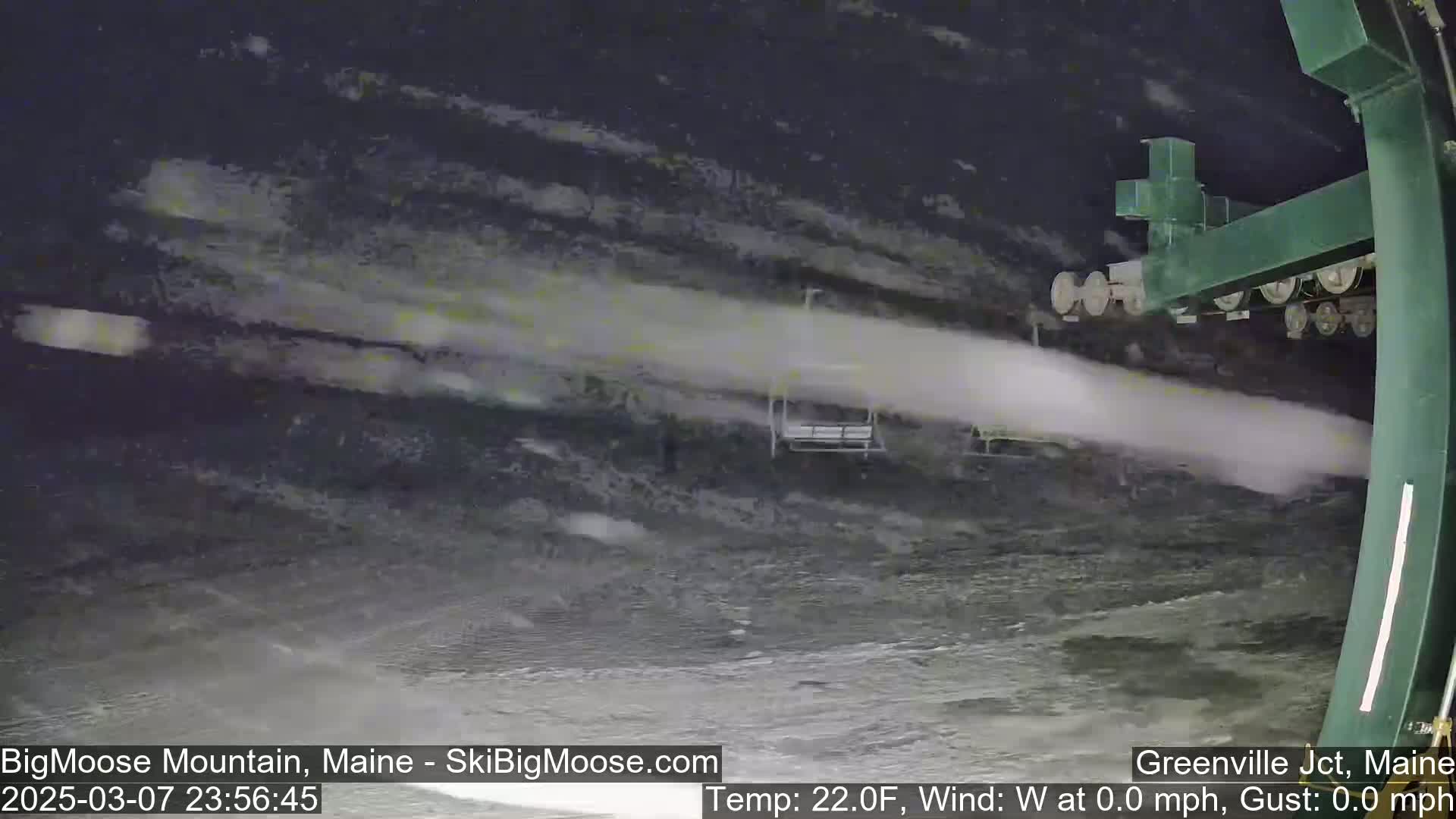 A chairlift ascends a snowy slope at night, with a plume of snow blowing from the lift mechanism.