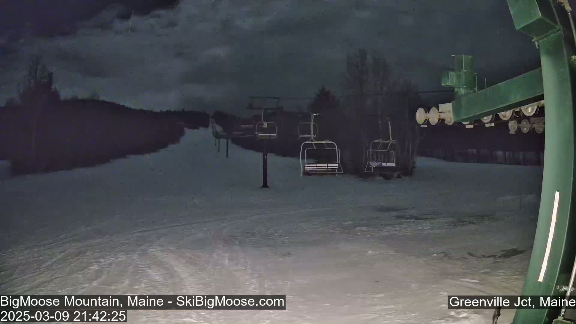A nighttime view of a snow-covered ski slope with empty chairlifts under a cloudy sky.