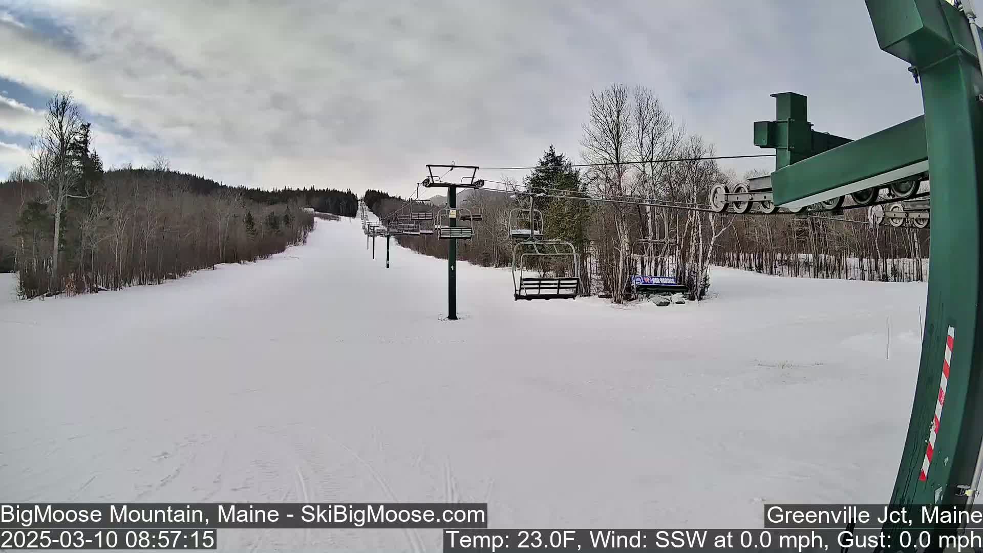 A snow-covered ski slope with a chairlift and bare trees under a cloudy sky.