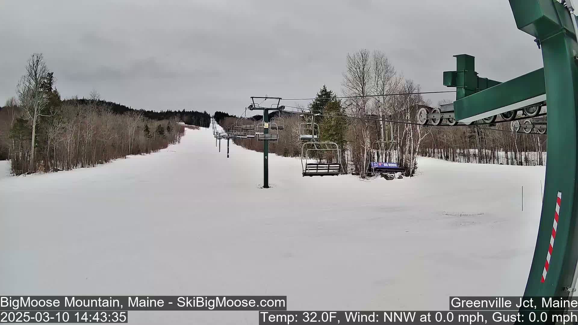 A snow-covered ski slope with a chairlift and sparsely wooded areas under an overcast sky.