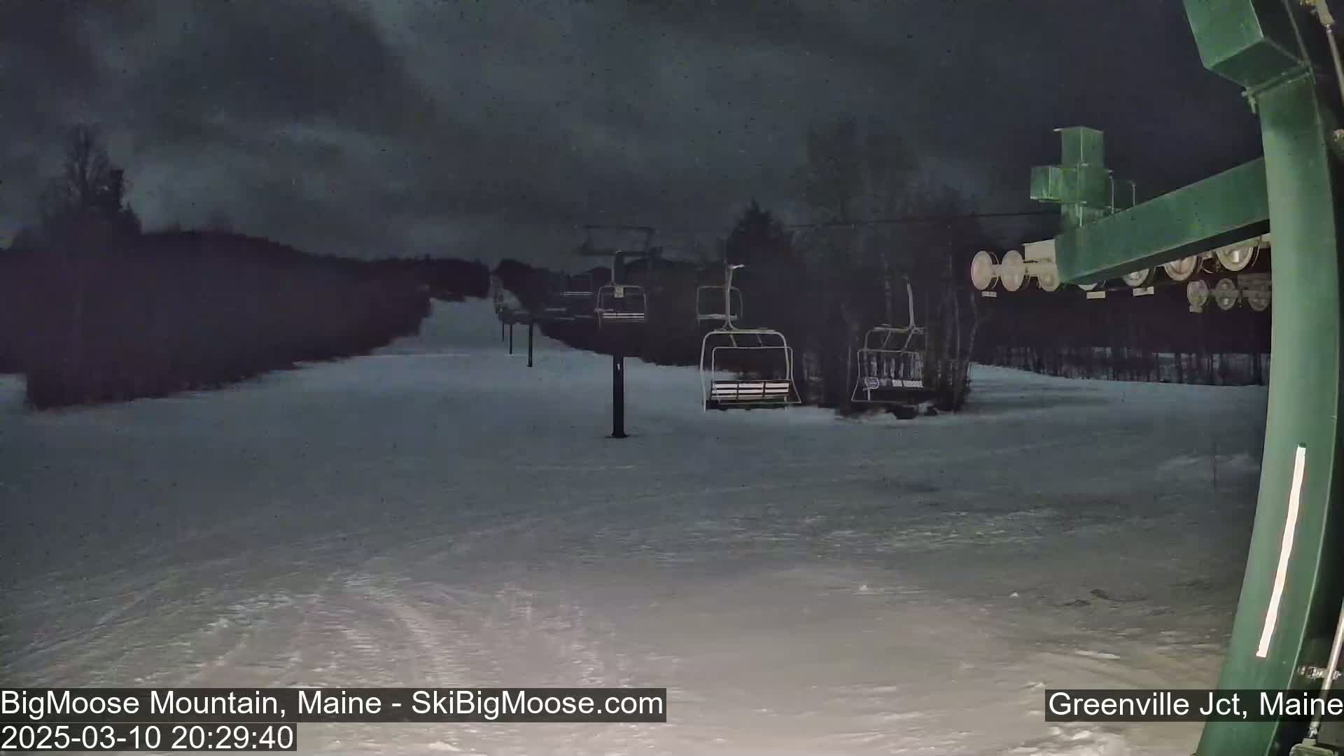 A snow-covered ski slope at night under a dark, cloudy sky shows several empty chairlifts.