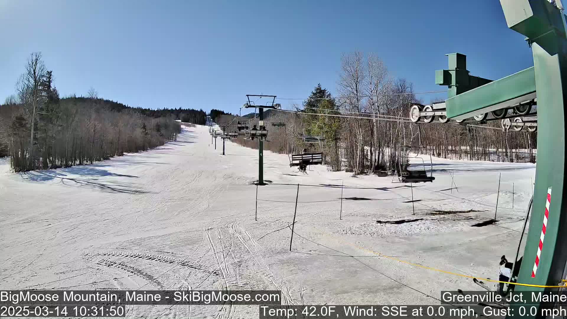 A snow-covered ski slope with a chairlift and surrounding trees under sunny skies.