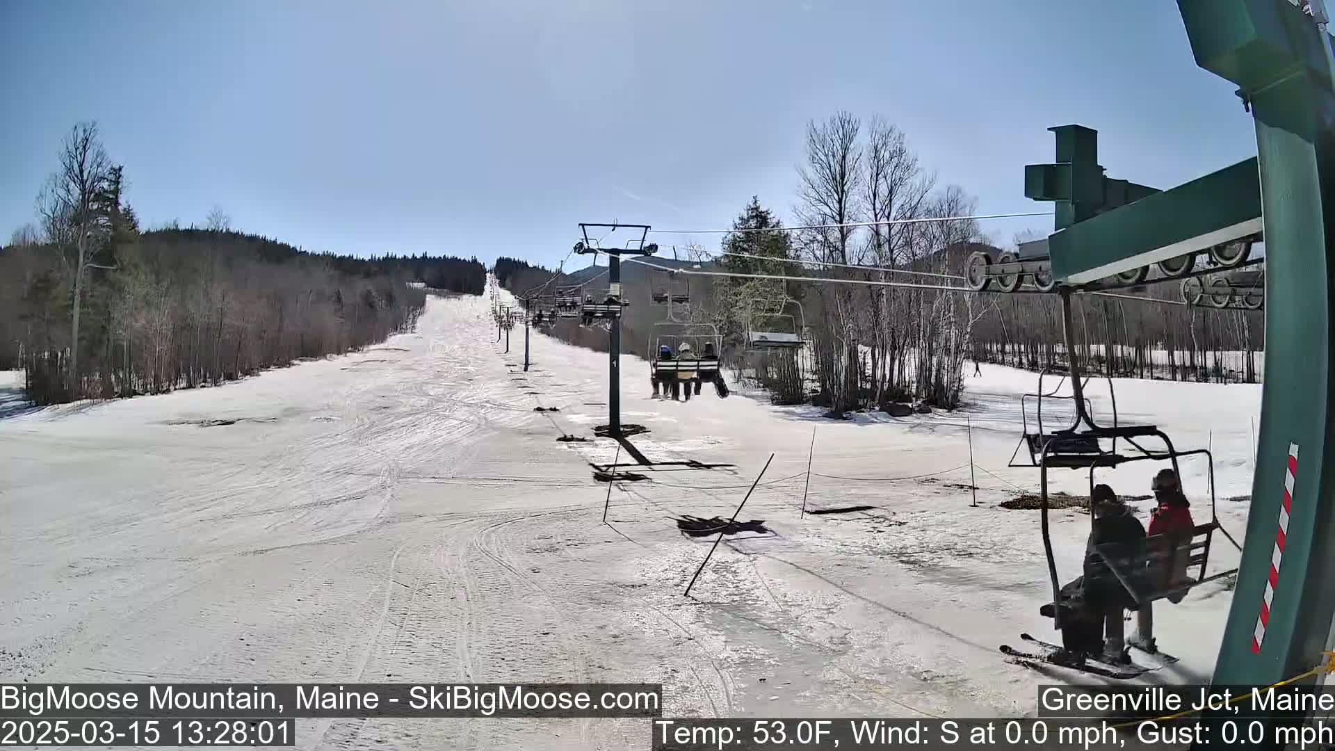 A sunny day at a ski resort shows a chairlift carrying skiers up a snow-covered slope, with bare trees lining the sides.