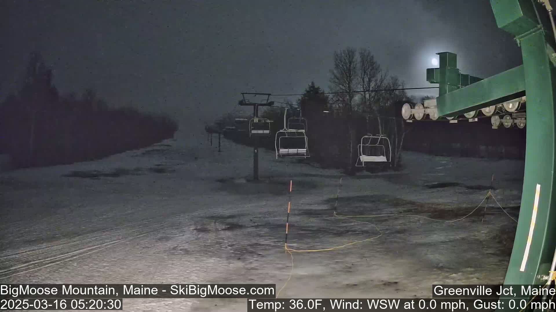 A nighttime view of a ski lift with empty chairs and a snowy, dark landscape under a dim moon.