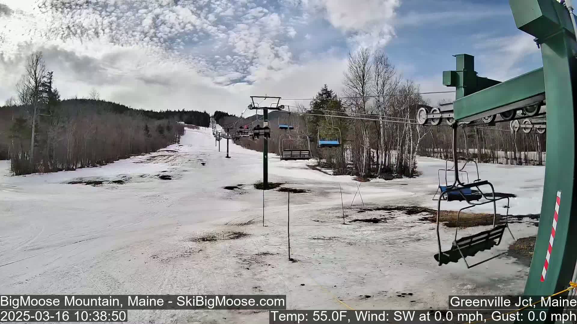 A mostly cloudy day at a ski resort shows a snow-covered ski slope with several empty chairlifts and trees in the background.
