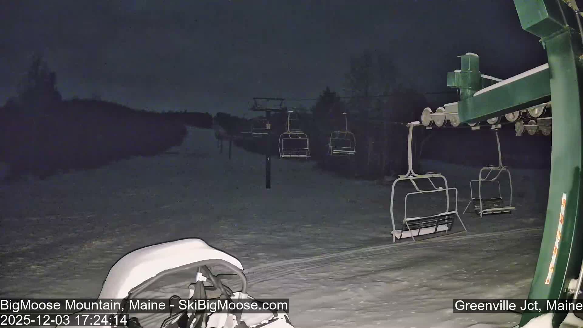 A clear, cold winter night view of a snowy ski slope with empty ski lift chairs ascending the track, and part of a snow-covered lift mechanism in the foreground.