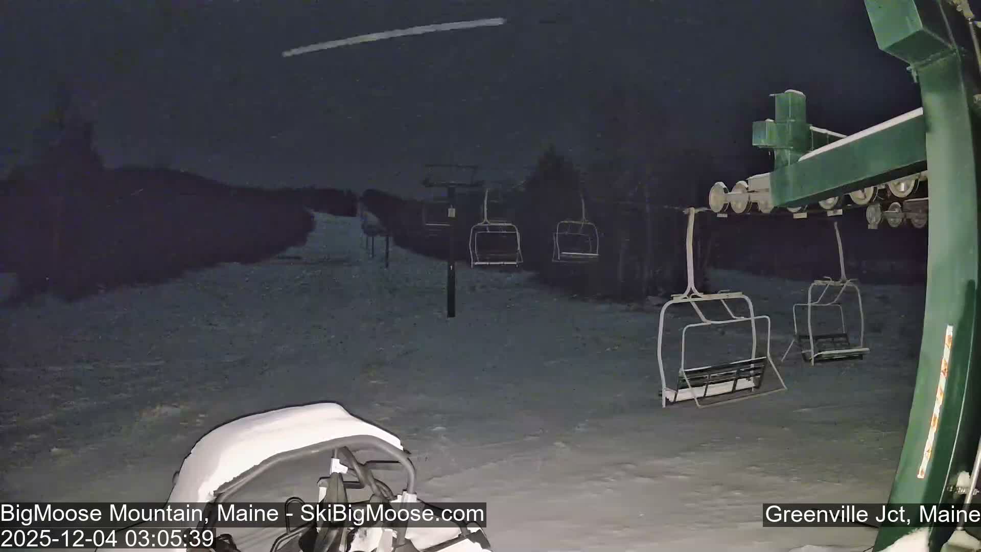 A snowy ski slope is seen at night with multiple empty ski lift chairs and part of a snow-covered vehicle in the foreground, under a dark sky with light snow falling.