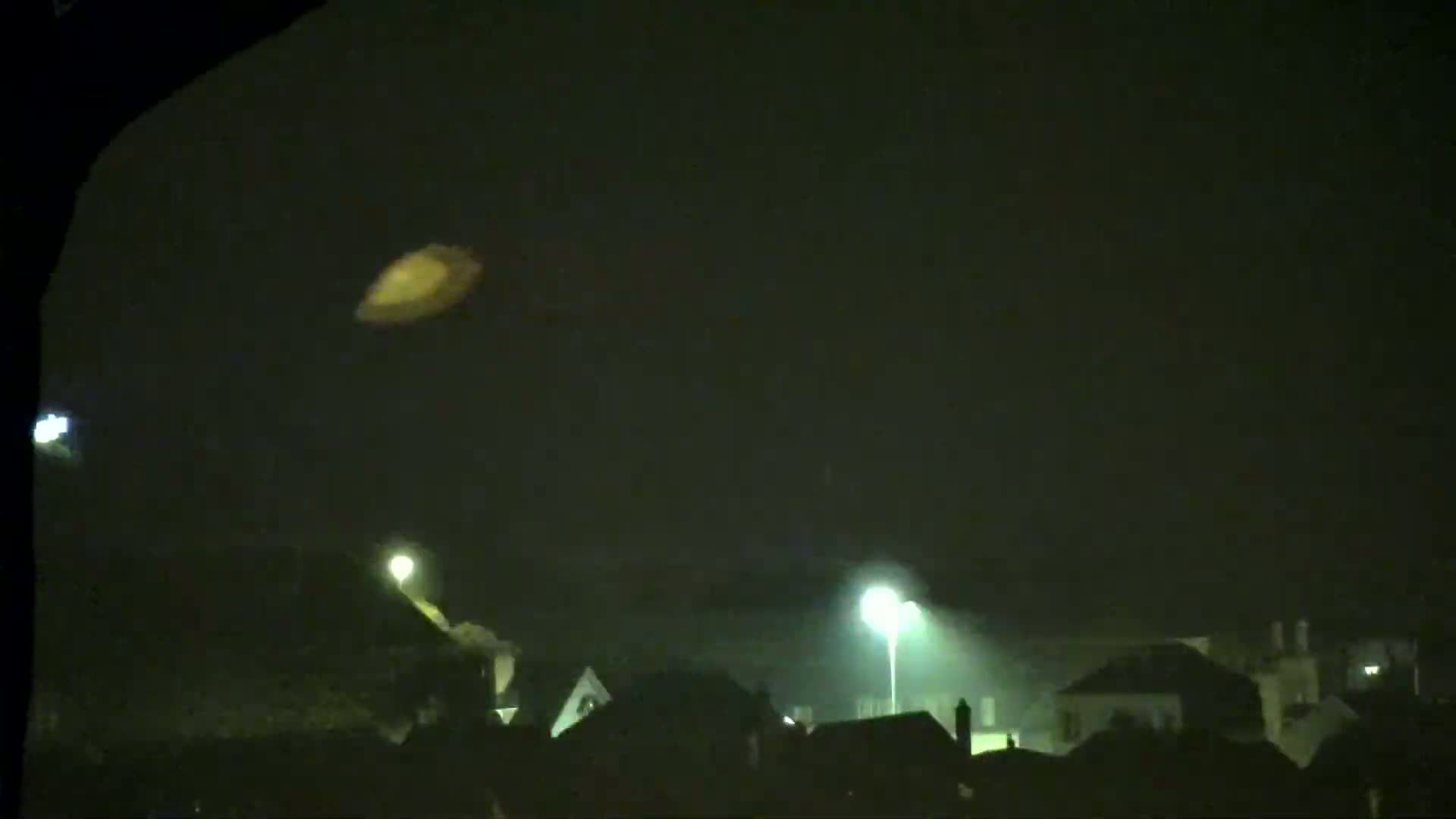 A blurry, yellowish-orange object is seen in the night sky above a foggy town illuminated by streetlights.