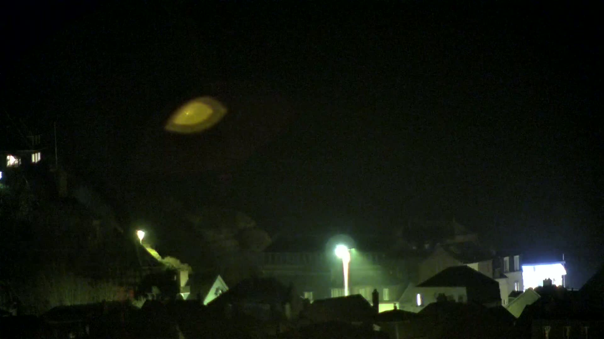A blurry, yellowish-white light hovers in a dark, nighttime sky above a town with dimly lit houses.