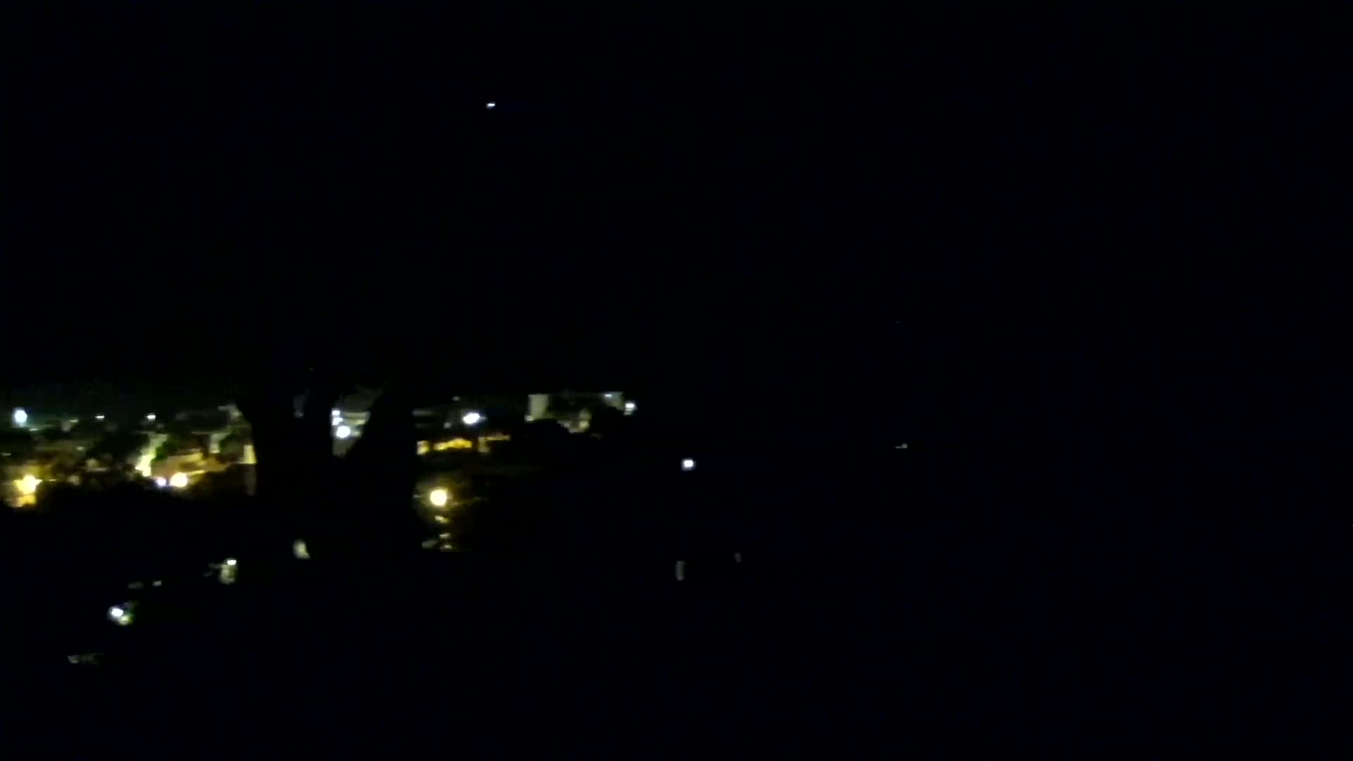 A nighttime view shows a distant town's lights obscured by a dark silhouette in the foreground under a clear, dark sky.