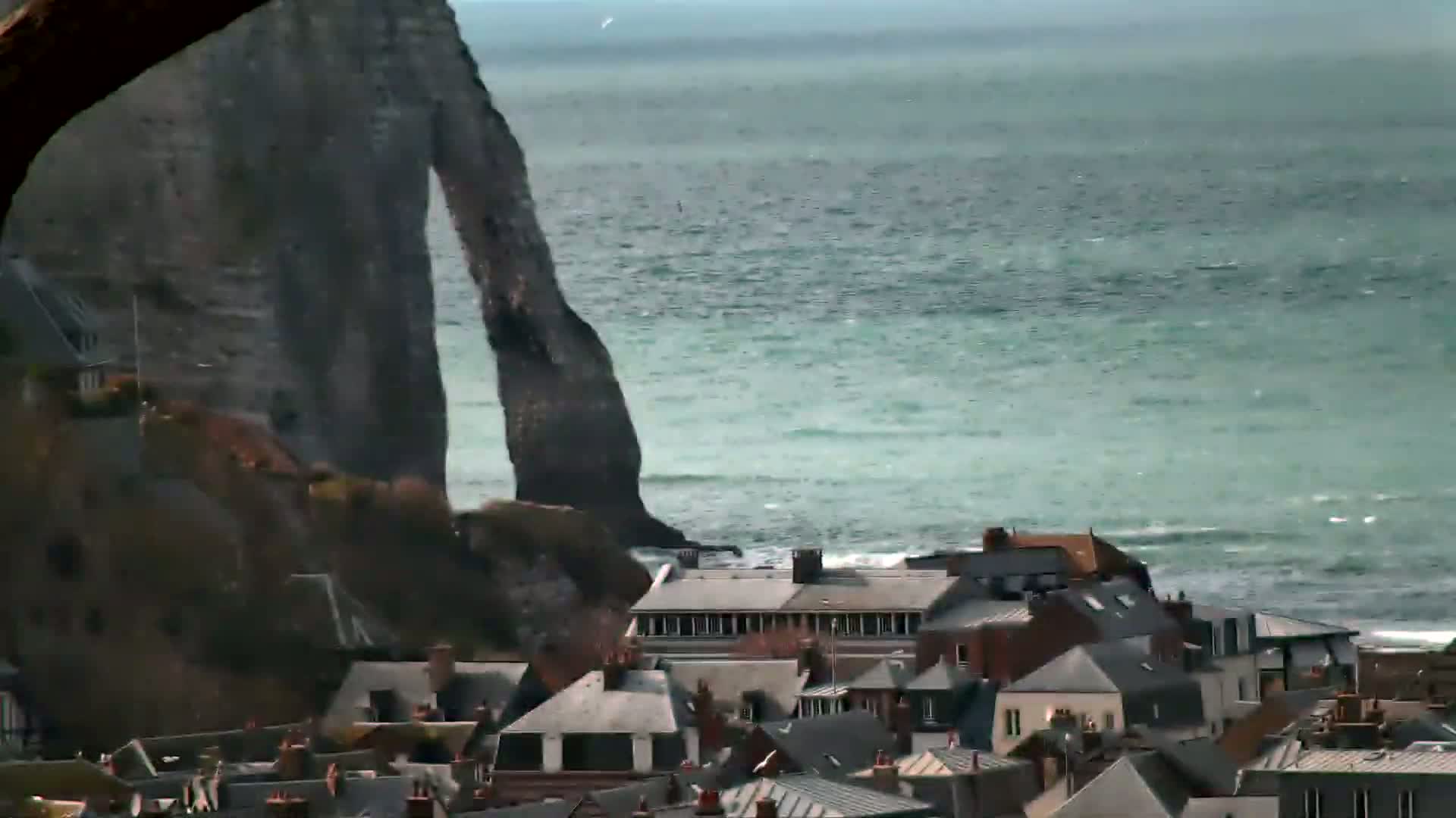 A coastal town with grey-roofed houses sits below a large, dark rock formation, overlooking a teal sea with small waves under a somewhat overcast sky.