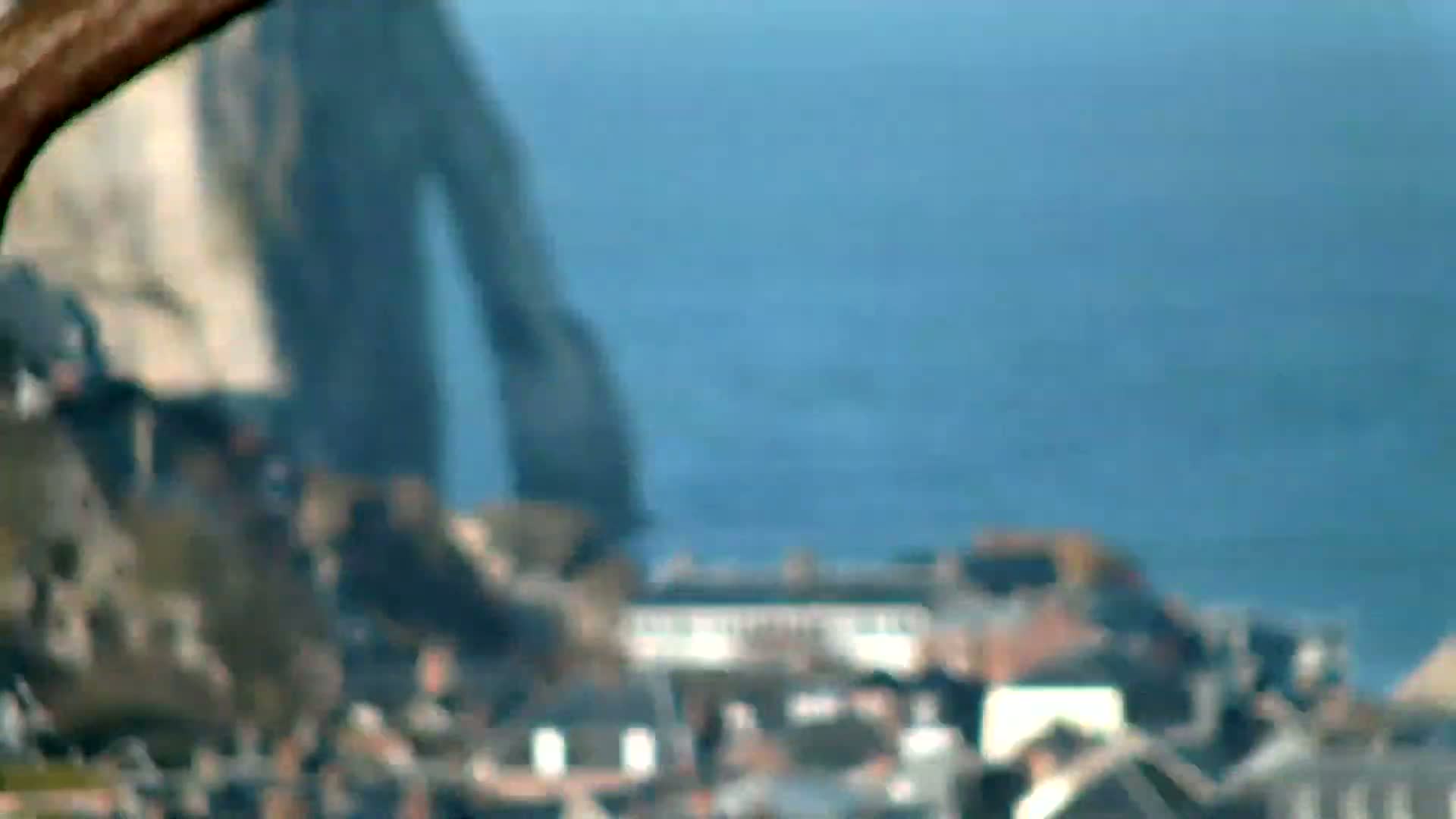 A blurry out-of-focus image shows a large, dark, indistinct object in the background, possibly a cliff face, overlooking a coastal town on a clear day.