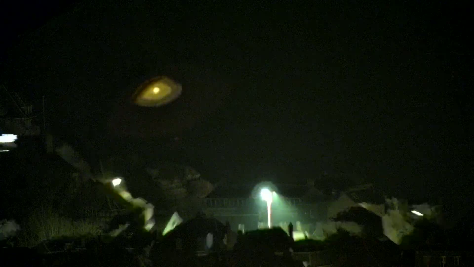 A blurry, illuminated object hangs in the night sky above a dimly lit town shrouded in fog or mist.