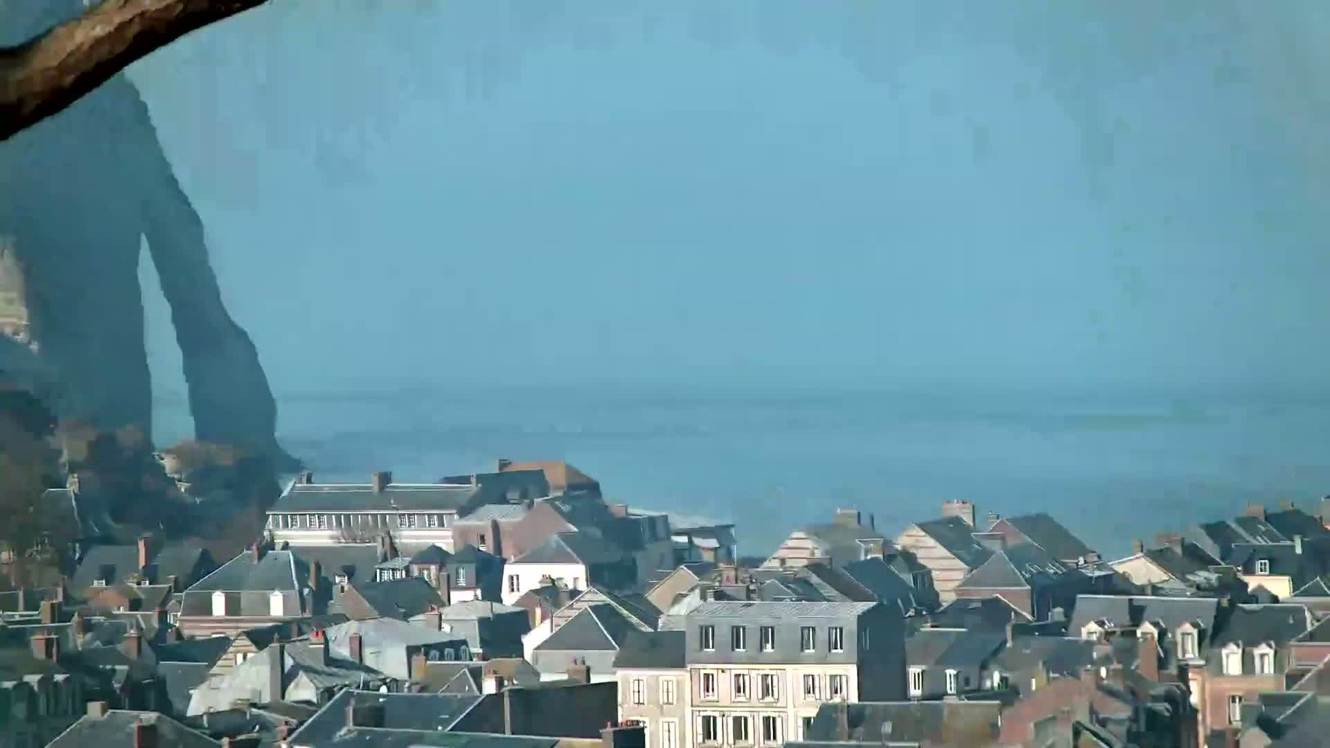 A coastal town with dark-grey slate roofs stretches towards a calm, light-blue sea under a clear sky, with a large rock formation visible to the left.