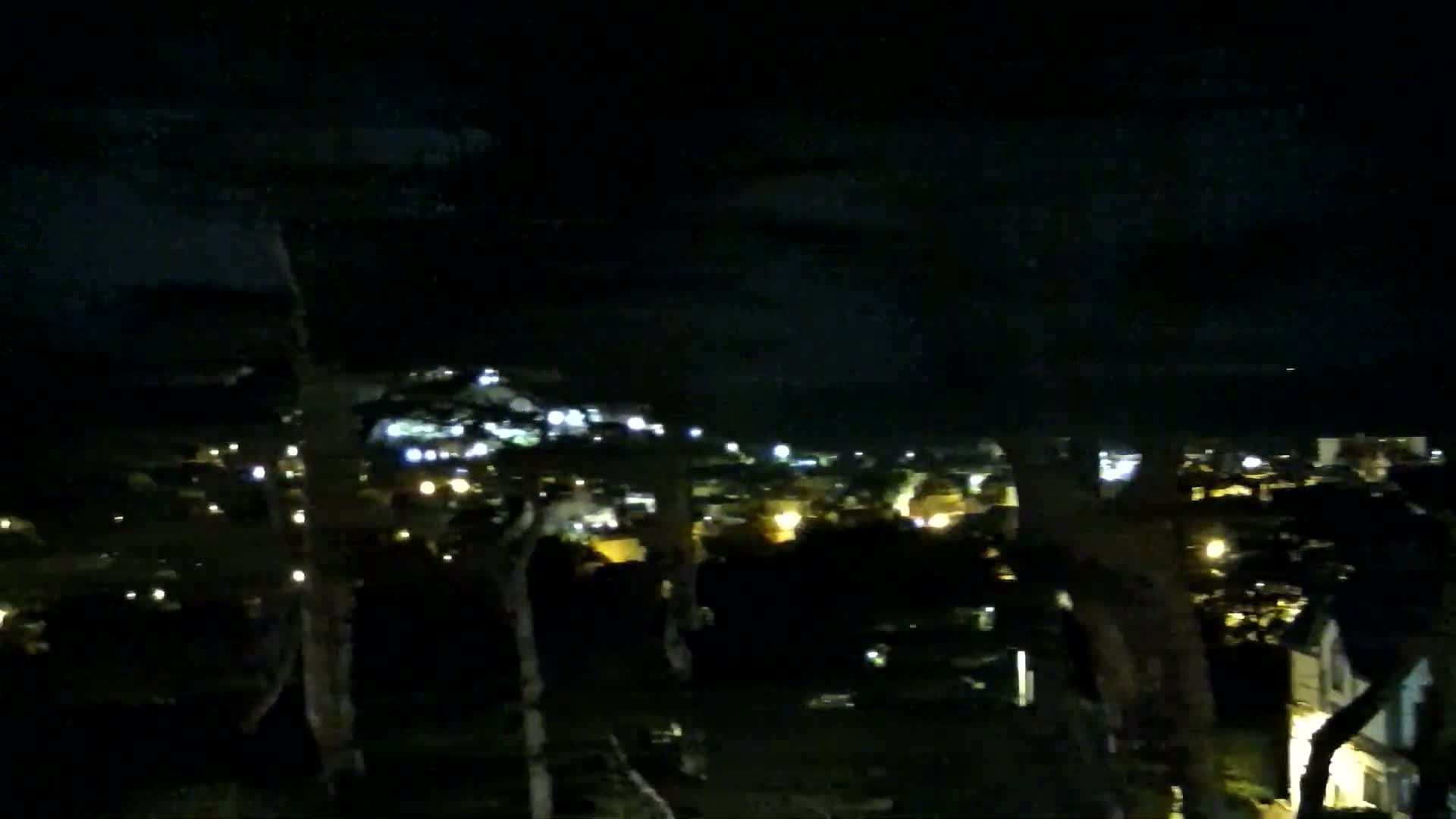 A nighttime view shows a dimly lit town nestled among dark trees under a cloudy sky.