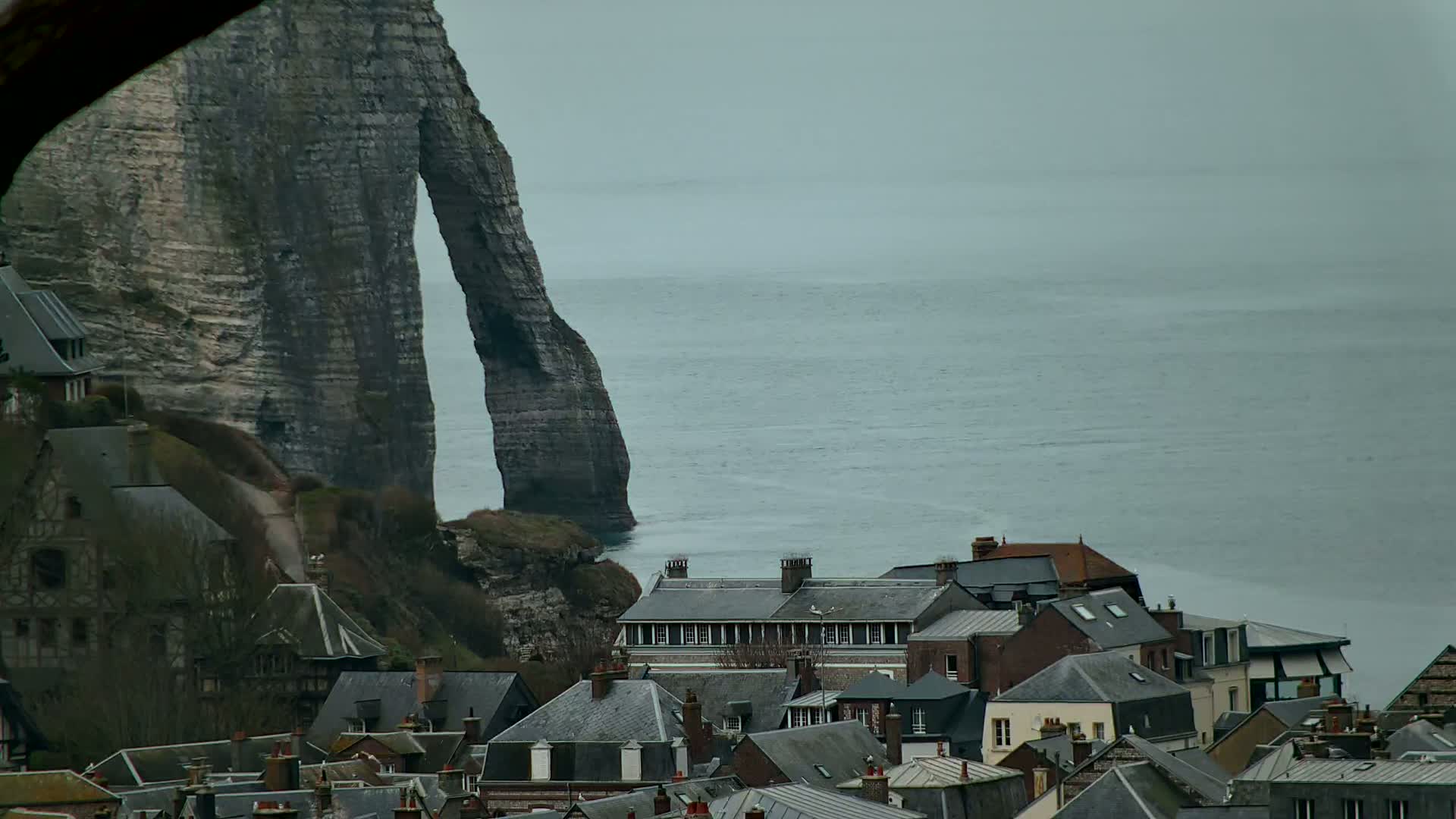 A coastal village with dark gray slate roofs nestles beneath a dramatic natural rock arch overlooking a calm, gray sea under an overcast sky.