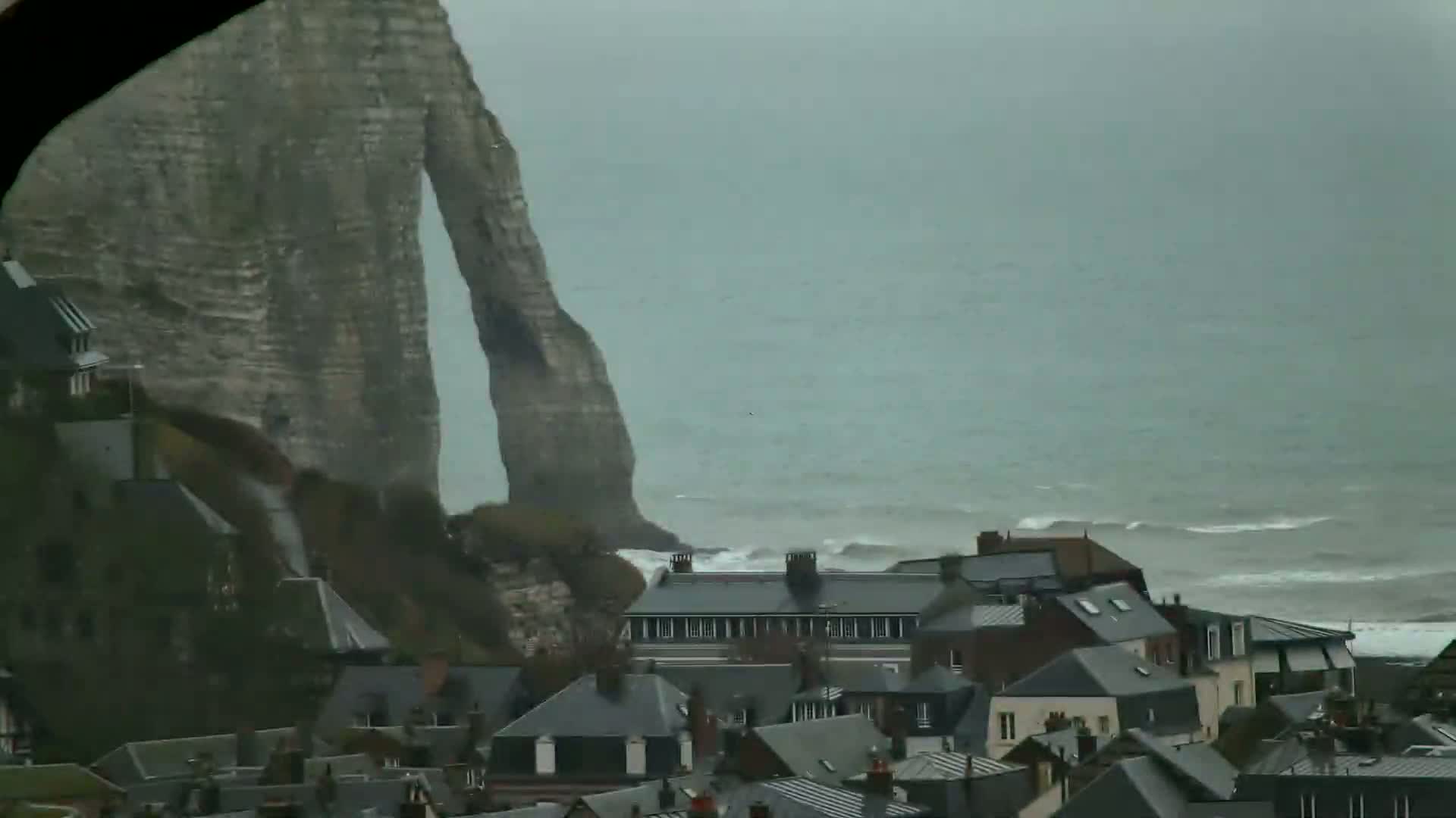 A large, imposing cliff overlooks a town of gray-roofed houses beside a choppy sea on an overcast day.