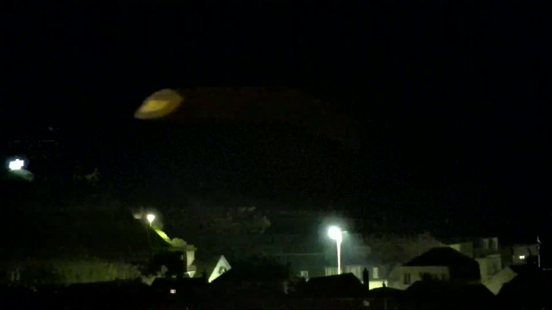 A faintly illuminated, oblong shape hovers in the night sky above a dimly lit residential area.