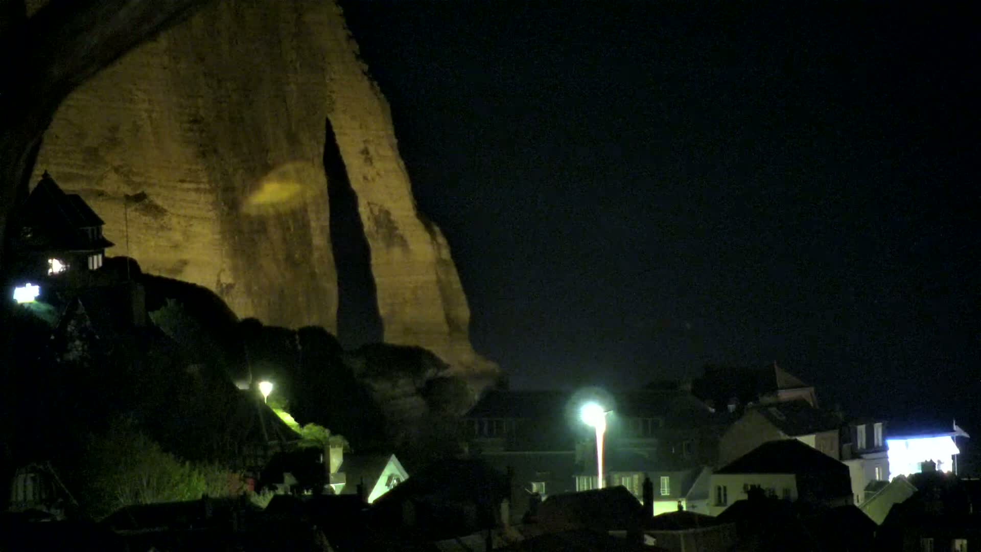 A large, illuminated cliff looms over a nighttime village of houses lit by streetlights.