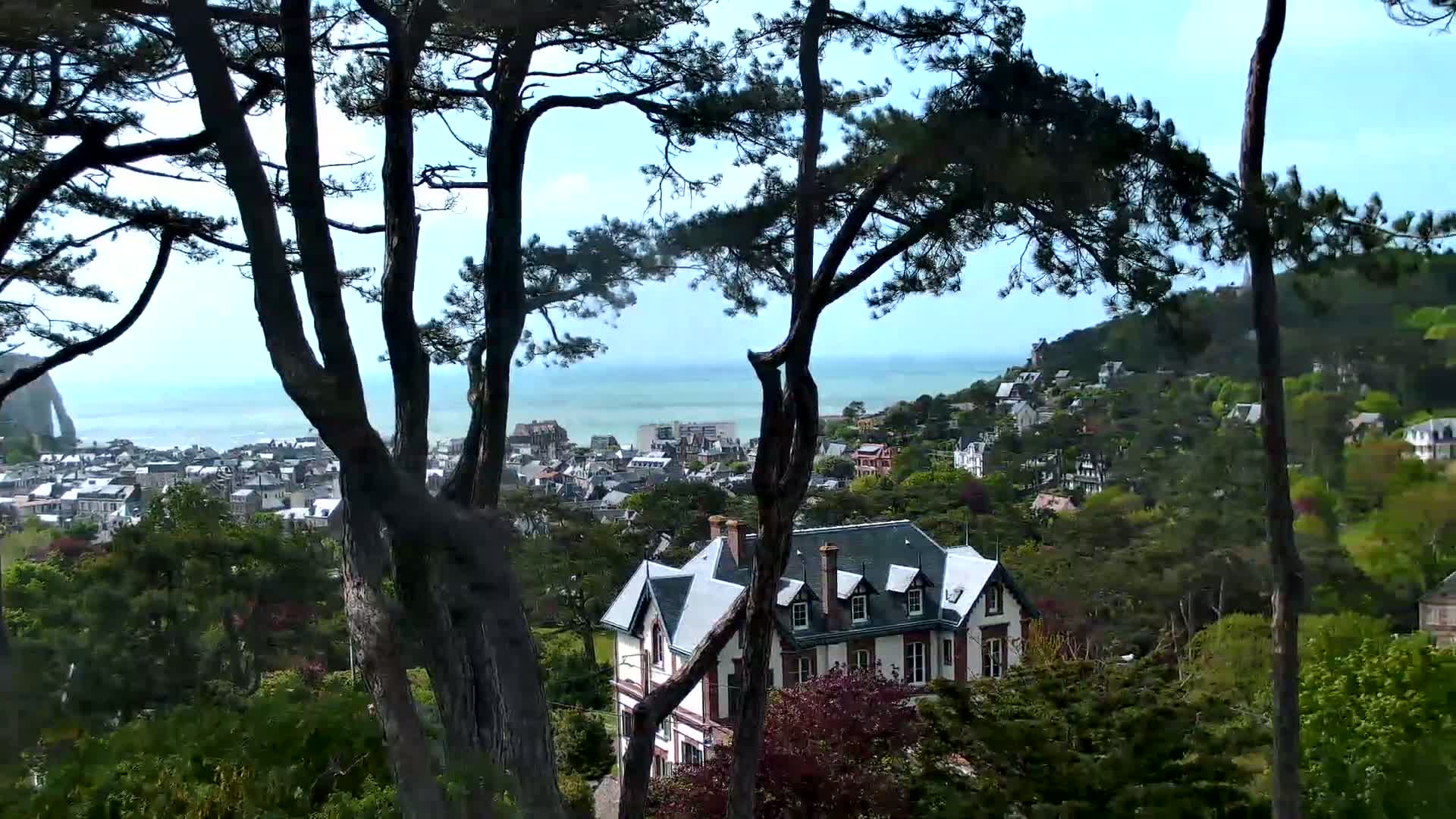 A large house sits nestled among lush green trees overlooking a coastal town and a calm ocean under a mostly clear sky.