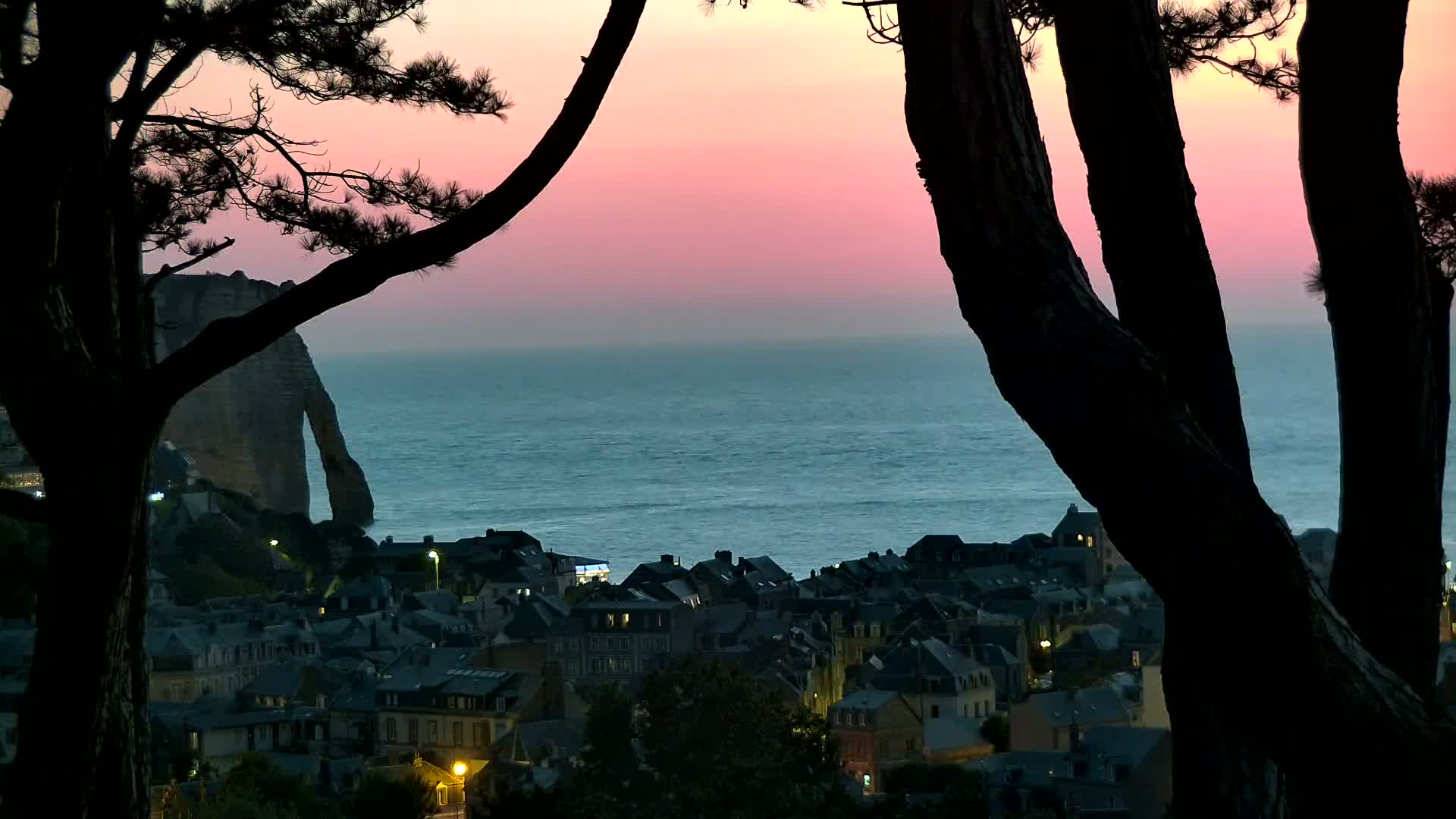 Silhouetted trees frame a view of a coastal town at dusk, with a pink sky over a calm ocean and a large rock formation visible in the distance.